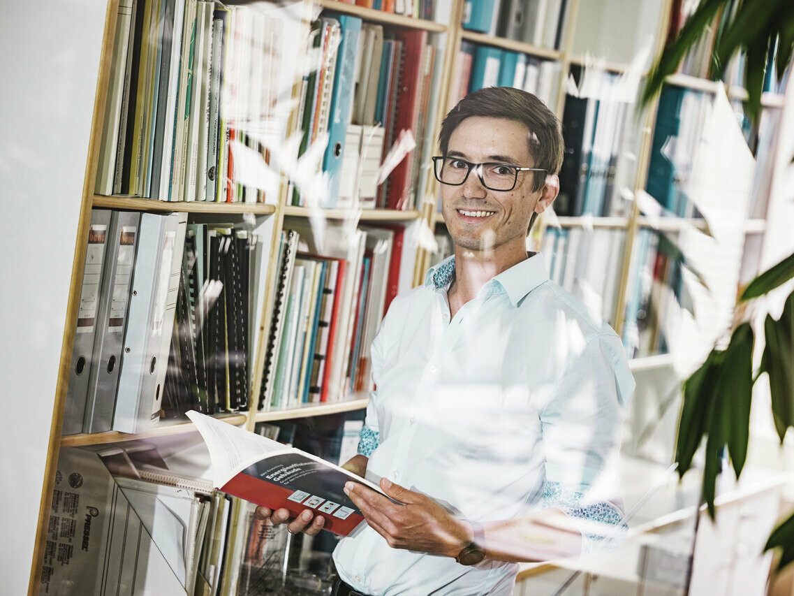 Architekt Christof Künz lächelnd  in einem hellen Hemd steht in einem Büro vor einem Bücherregal, das mit Ordnern und Büchern gefüllt ist. Er hält eine geöffnete Broschüre in den Händen. Die Szene wird durch eine Pflanze im Vordergrund und die Spiegelung des Bürointerieurs auf einer Glasscheibe belebt, was eine freundliche und wissensreiche Atmosphäre vermittelt.