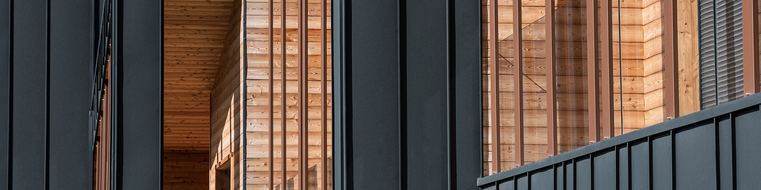 Nahaufnahme der Fassade eines modernen Gebäudes, die eine elegante Symbiose aus dunkelgrauen Prefalz Bahnen und warm getönten, natürlich gemaserten Holzbalken zeigt, die Struktur und Tiefe verleihen und durch die Sonneneinstrahlung beleuchtet werden.
