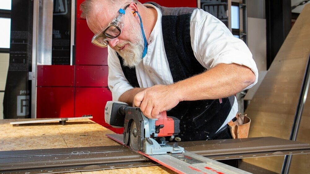 Das Bild zeigt einen Mann, der an einer Werkbank arbeitet. Er schneidet mit einer Sägemaschine das PREFA Siding-Fassadenelement zu. Er ist dabei sehr fokussiert, die Detailgenauigkeit seiner Arbeit kommt zum Vorschein.