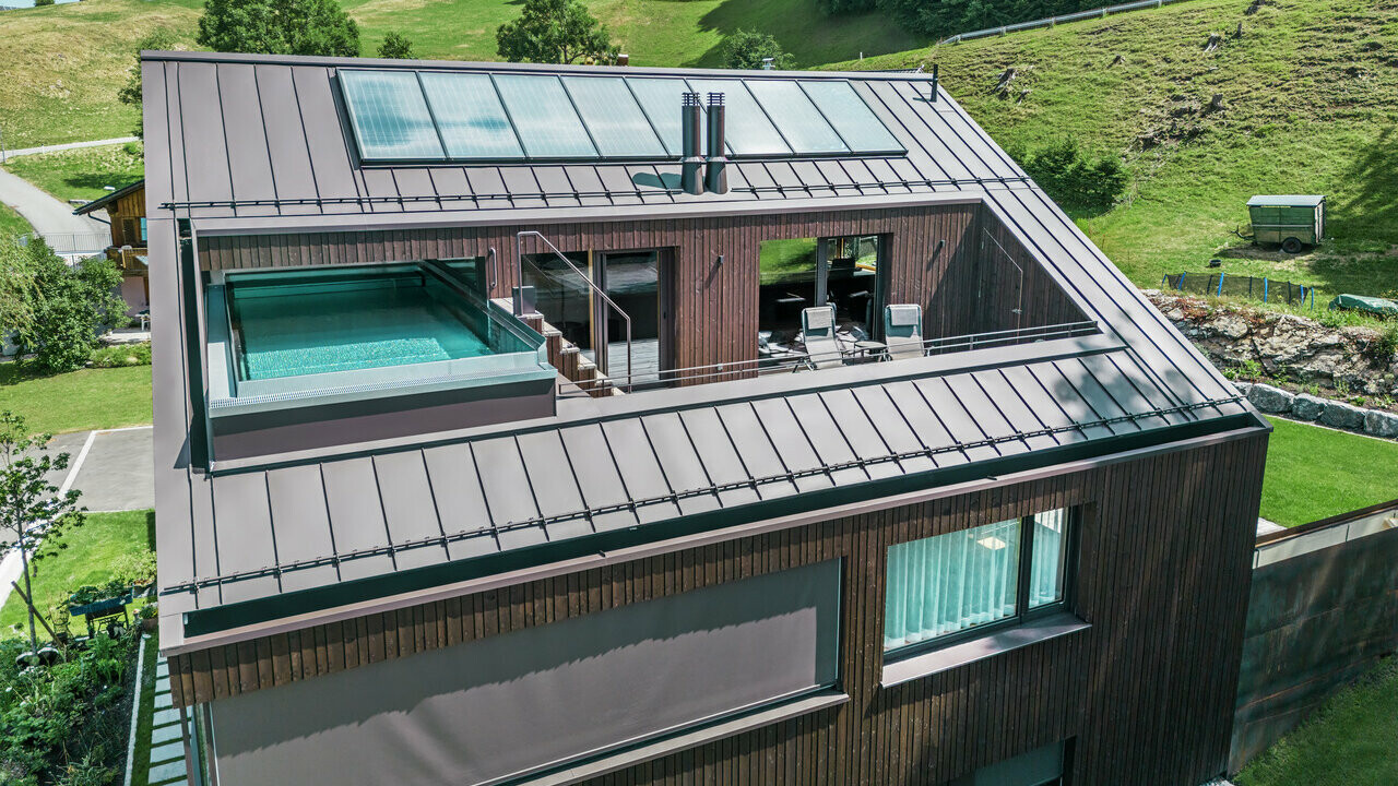 Ansicht eines modernen Mehrfamilienhauses mit einem PREFA Dach in Prefalz-Technik, ausgeführt in der Farbe Nussbraun. Das Bild zeigt auch eine Terrasse mit einem Pool, umgeben von einer Holzverkleidung und Glasgeländern.