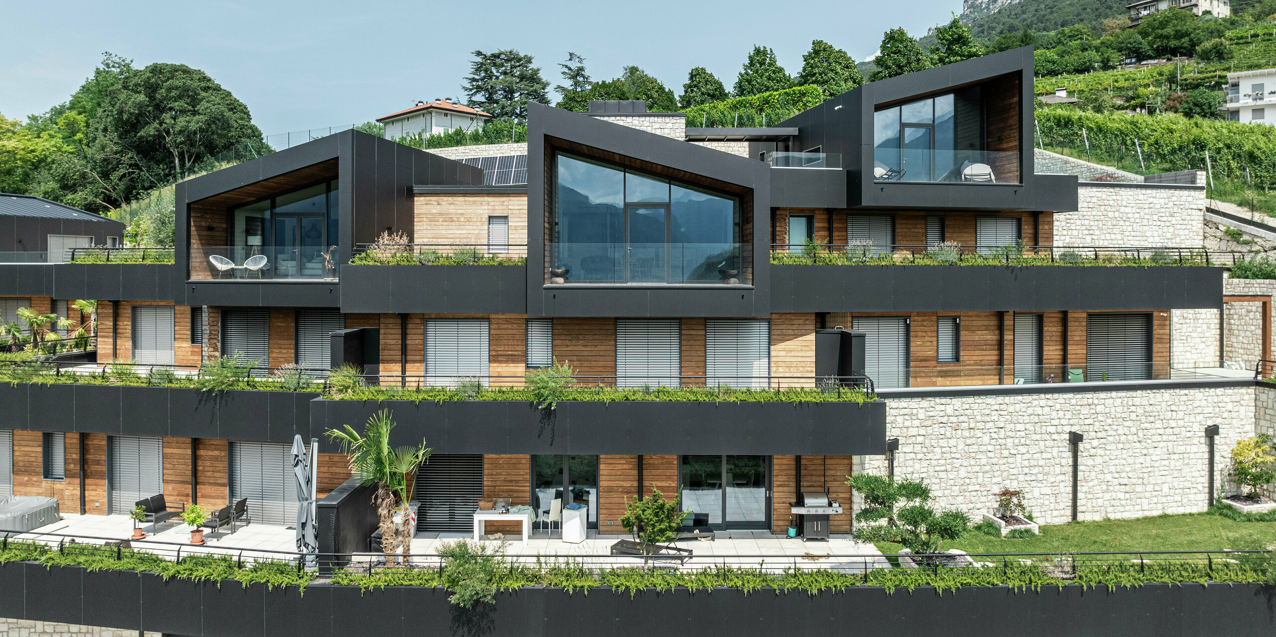 Eleganz und Funktionalität in Perfektion: Die markante Architektur dieser Wohnanlage zeichnet sich durch moderne Giebelformen, große Glasflächen und einen harmonischen Materialmix aus. Die Fassade aus PREFA Aluminium Verbundplatten in P.10 Schwarz sorgt für eine widerstandsfähige und wartungsfreie Gebäudehülle, während das PREFALZ Dach optimalen Schutz bietet. Holz und Naturstein setzen warme Akzente und fügen sich nahtlos in die grüne Umgebung ein. Begrünte Terrassen und großzügige Wohnbereiche schaffen ein einzigartiges Wohngefühl mit höchstem Komfort und Panoramablick.