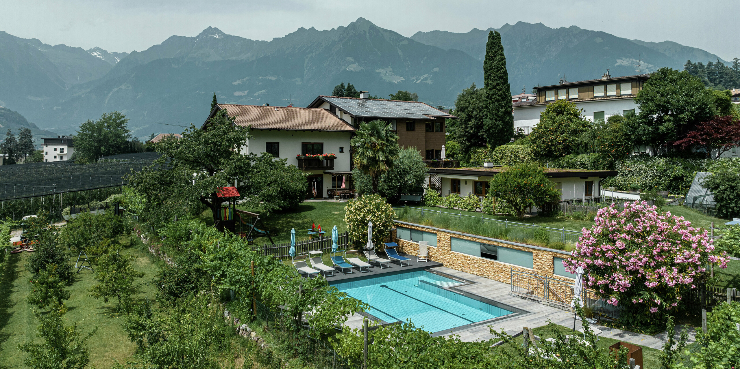 Ferienwohnungen Prechtlhof in Meran, Südtirol, umgeben von Obstgärten und Bergpanorama. Die sanierte Fassade mit PREFA Sidings in Walnussbraun fügt sich harmonisch in die Landschaft ein. Im Vordergrund lädt ein Poolbereich mit Liegestühlen und bunten Blumen zur Entspannung ein.