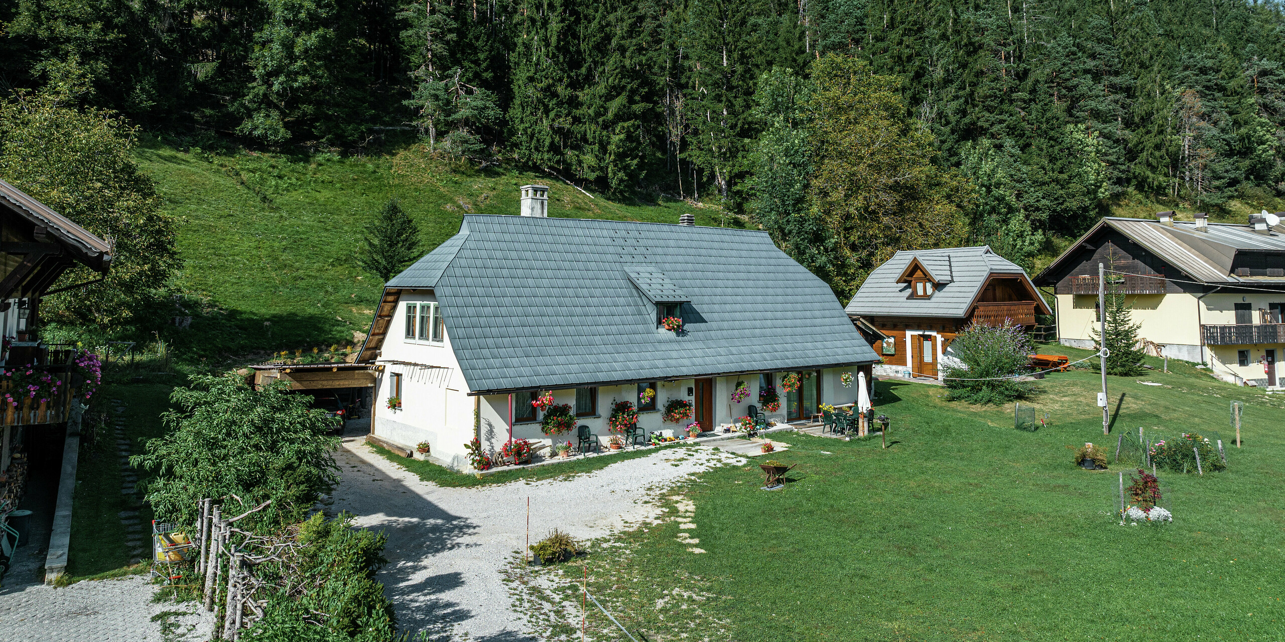 Luftaufnahme eines idyllischen Einfamilienhauses in Tarvis, Italien, das seit 1972 von langlebigen PREFA Dachplatten geschützt wird. Das großflächige, witterungsbeständige Aluminiumdach fügt sich harmonisch in die grüne Alpenlandschaft ein. Die liebevoll mit Blumen geschmückte Fassade und die gemütliche Terrasse verleihen dem Haus eine einladende Atmosphäre. Umgeben von traditionellen Häusern mit Holzfassaden und dichtem Nadelwald bietet das Anwesen eine perfekte Mischung aus Naturverbundenheit und architektonischer Beständigkeit.