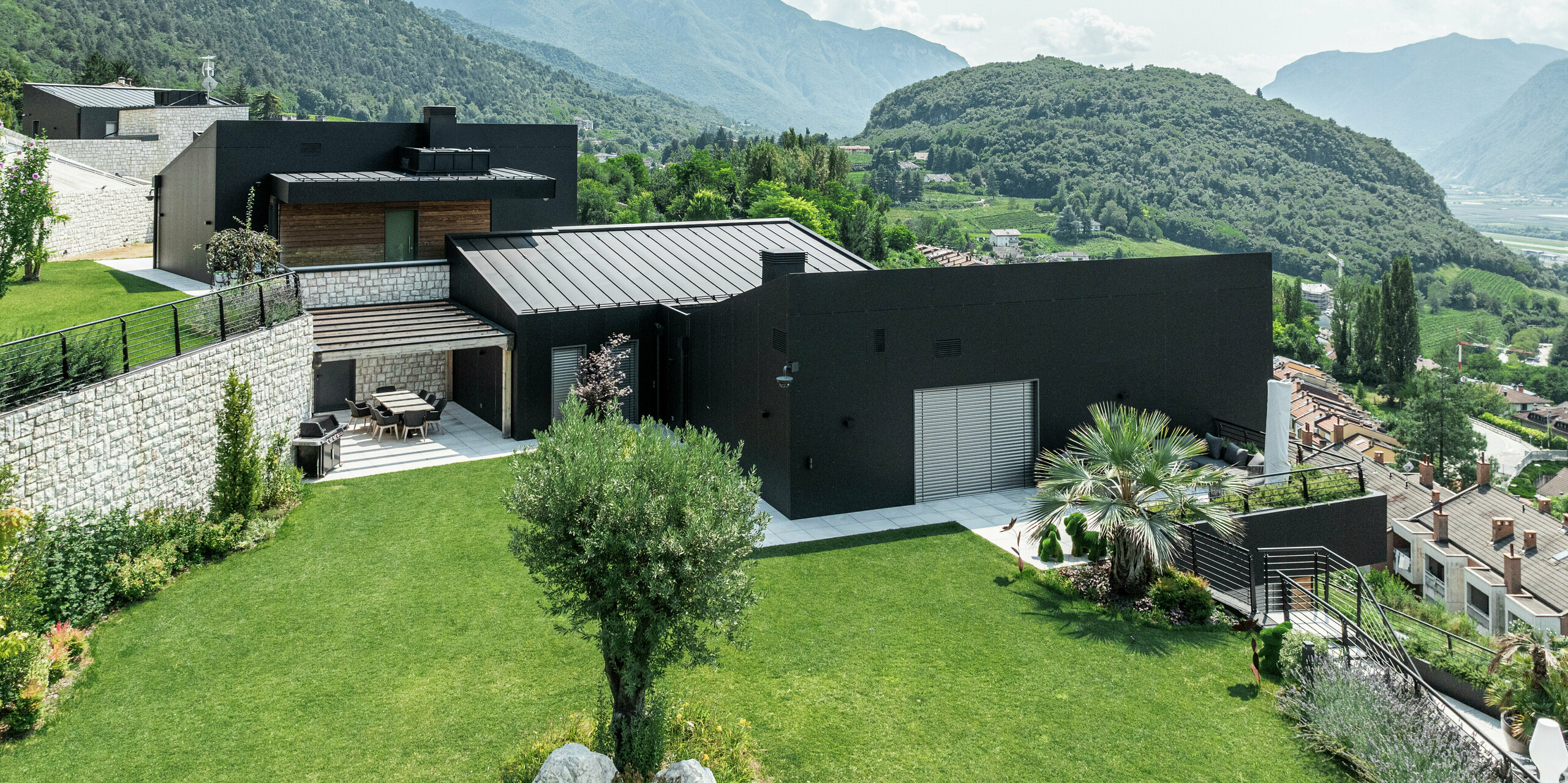 Moderne Wohnhausanlage in Hanglage mit atemberaubendem Blick auf die Trienter Berglandschaft. Die Fassade aus PREFA Aluminium Verbundplatten in P.10 Schwarz sorgt für eine elegante und witterungsbeständige Gebäudehülle. Das PREFALZ Dach in P.10 Schwarz rundet das minimalistische Design ab und bietet höchste Langlebigkeit. Eine gepflegte Gartenlandschaft mit Natursteinmauern fügt sich harmonisch in die Umgebung ein und unterstreicht den exklusiven Wohncharakter.