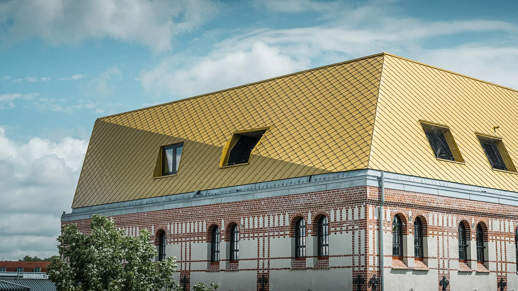 Das Dach mit den mayagoldenen Aluminiumrauten und einem Knick aus der Nähe, ein Teil der denkmalgeschützten Fassade ist sichtbar.