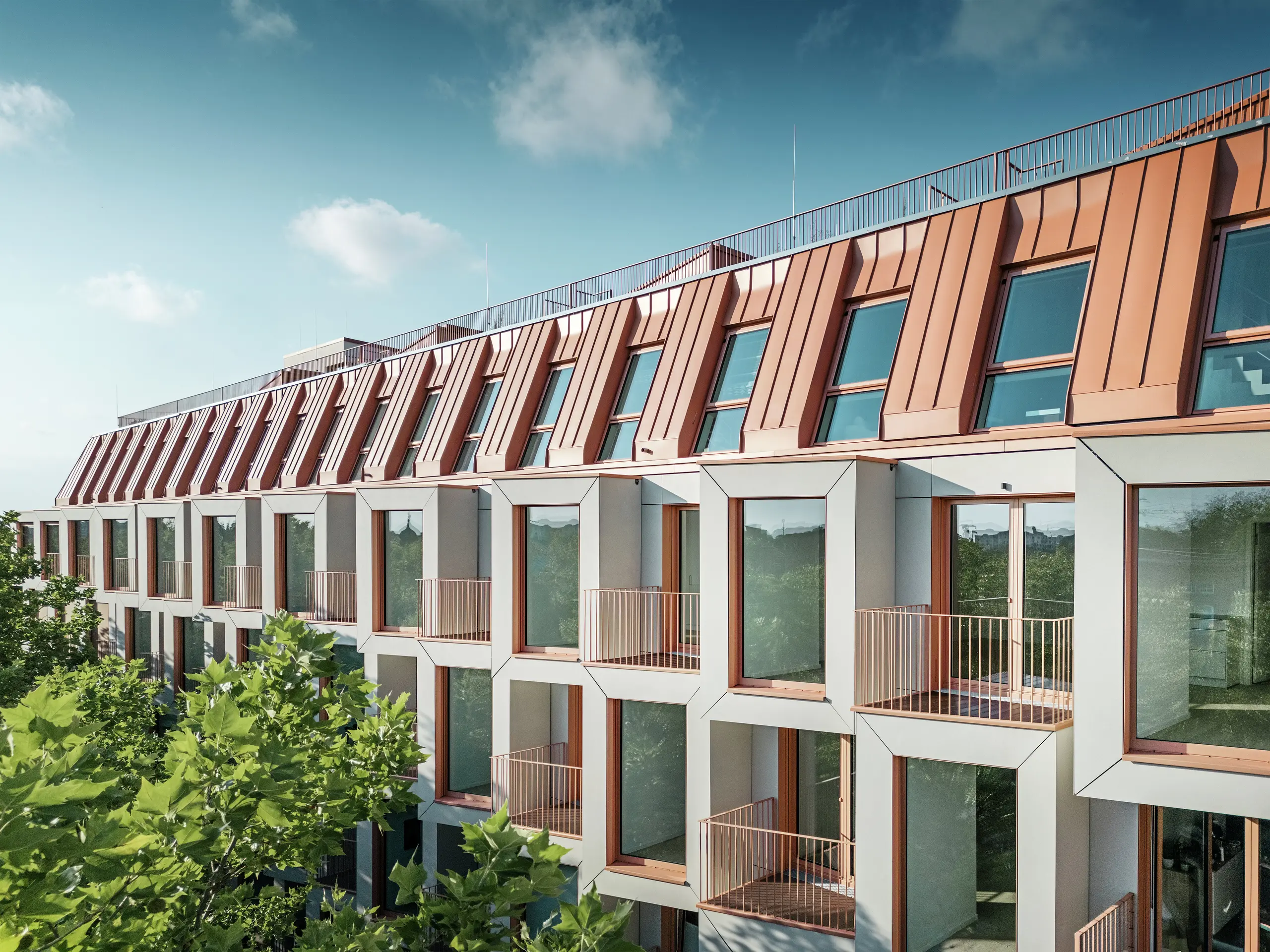 Blick auf die Fassade des Wohnprojekts Van B in München mit dem markanten PREFALZ-Aluminiumdach in der Sonderfarbe IGP Orange HDP von PREFA. Die moderne Dachkonstruktion vereint ästhetische Eleganz mit technischer Präzision und bietet dauerhaften Schutz vor Witterungseinflüssen. Kubische Erker mit großen Fensterflächen und sanft geschwungene Balkone schaffen ein harmonisches Zusammenspiel von Licht, Raum und Funktionalität – ideal für anspruchsvolle Wohnkonzepte im urbanen Raum.