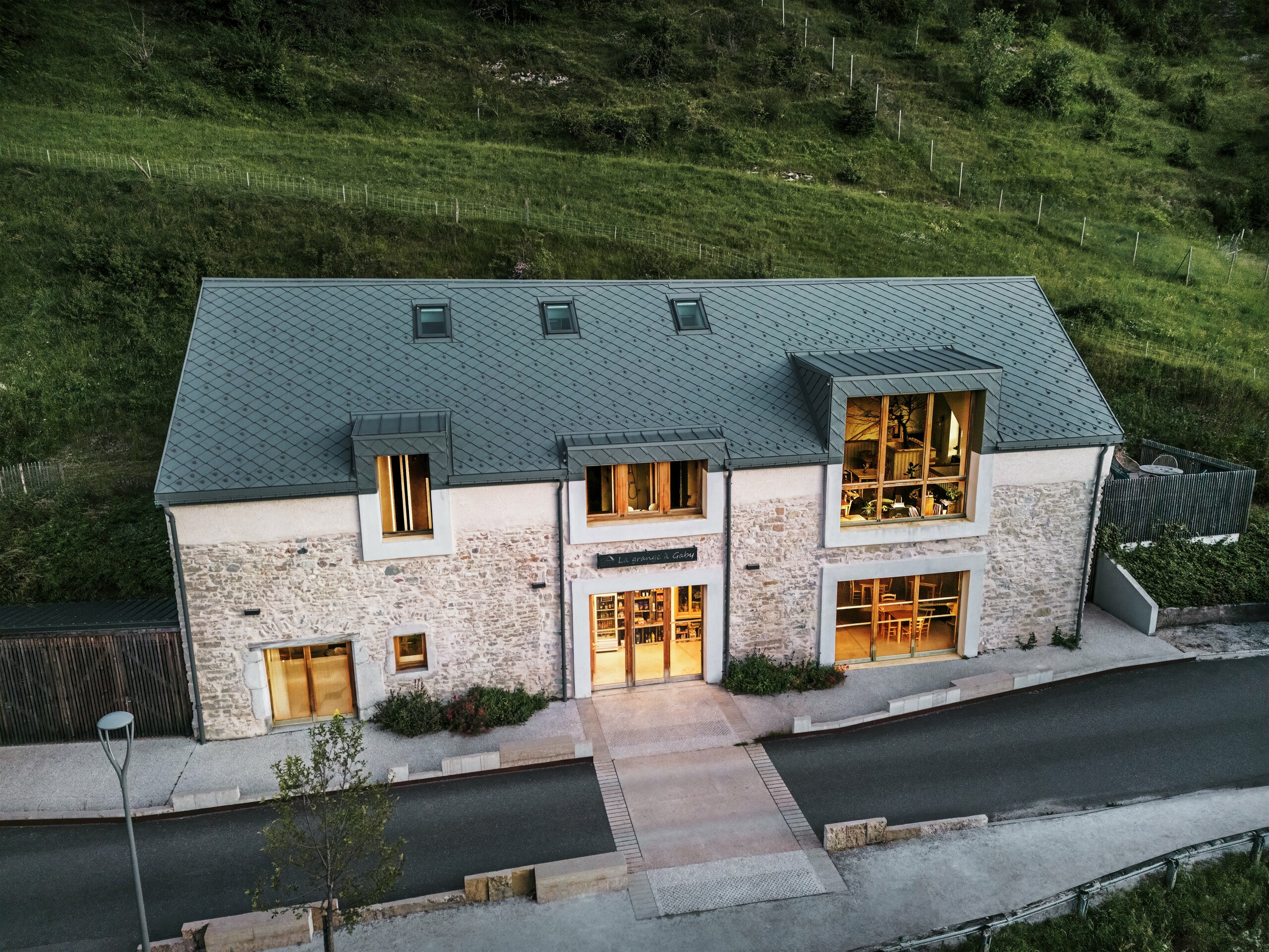 Abendansicht der restaurierten Scheune "La Grange à Gaby" in Engins, Frankreich. Das Gebäude kombiniert traditionelles Steinemauerwerk mit einem modernen PREFA Aluminiumdach aus 29 × 29 cm großen Rauten. Die großen Fenster und die beleuchteten Innenräume unterstreichen die einladende Atmosphäre des neuen kulturellen Zentrums.