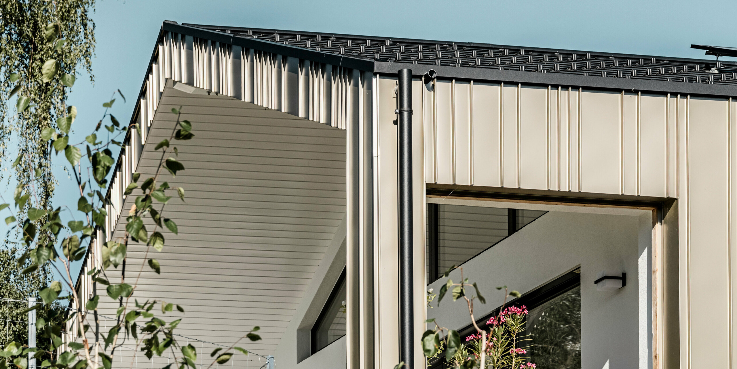 Moderne Architektur mit einer PREFA Aluminiumfassade in Lichtbronze. Abgerundet wird das Erscheinungsbild durch eine anthrazitfarbene Dachkante und eine hochwertige PREFA Dachentwässerung. Die vertikale Anordnung der Winkelstehfalzdeckung betont die Höhe des Gebäudes und verleiht ihm eine elegante Textur, während die harmonische Farbgebung sich nahtlos in die natürliche Umgebung einfügt. Dieses Bild zeigt die Beständigkeit und Ästhetik von PREFA Aluminiumprodukten, die für zeitgemäßes Bauen und innovatives Design stehen.