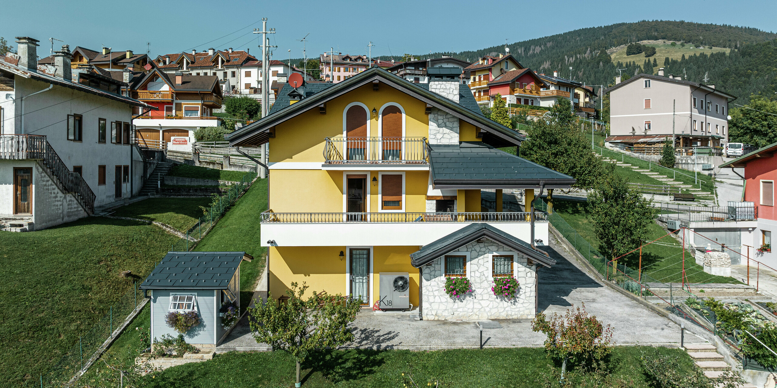 Einfamilienhaus in Gallio mit alpinem Charme und modernem PREFA Aluminiumdach in P.10 Dunkelgrau. Die langlebigen PREFA Dachplatten R.16 sorgen für optimalen Witterungsschutz und eine ansprechende Optik. Die gelbe Fassade an der Gebäuderückseite wurde mit Natursteinelementen und Holzakzenten kombiniert - eine perfekte Symbiose mit der grünen Umgebung. Abgerundet wird das hochwertige Blechdach durch die PREFA Dachentwässerung, die für eine zuverlässige Wasserableitung sorgt. Ein elegantes Dachsystem, das Funktionalität und Ästhetik vereint.