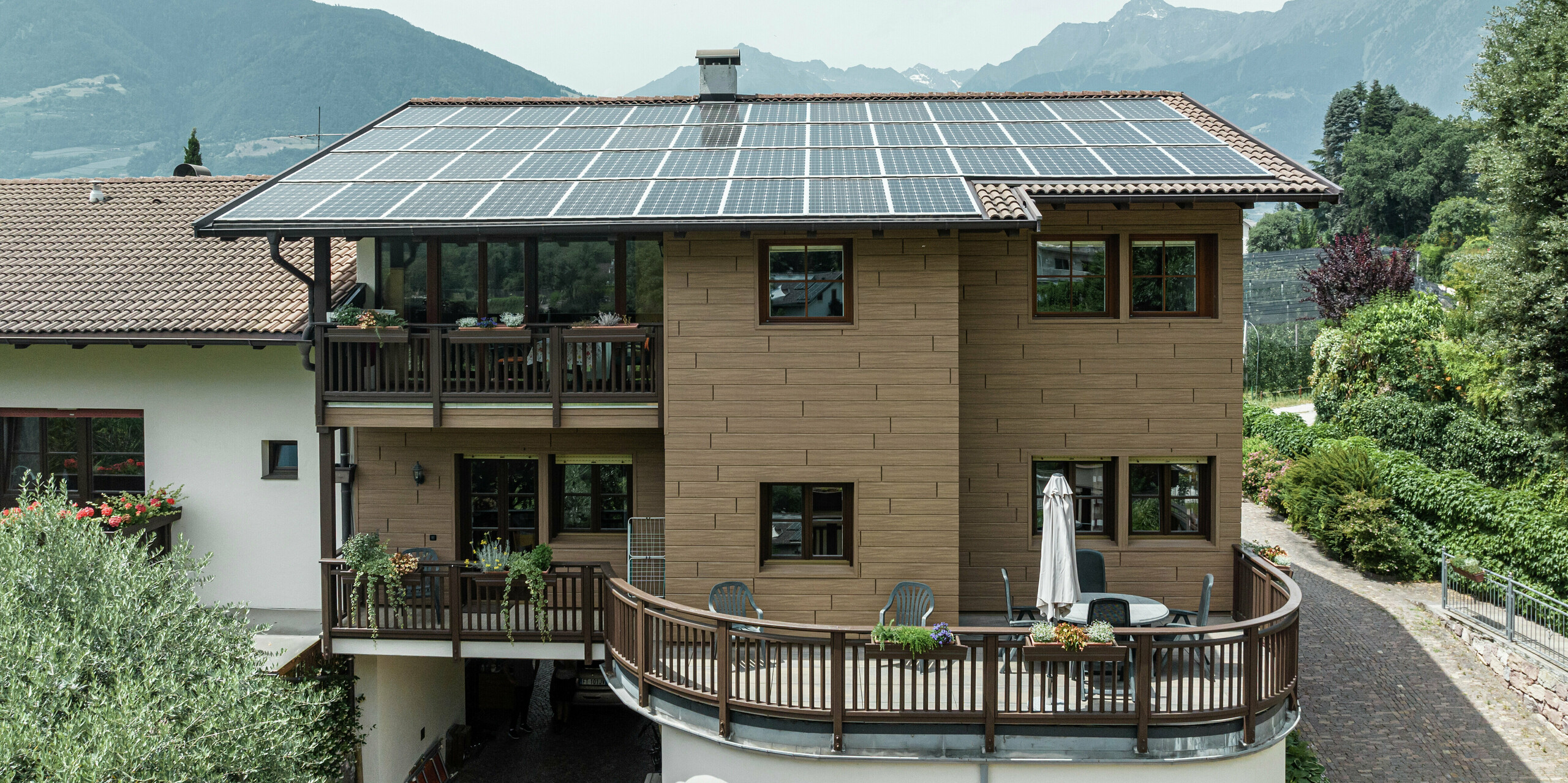 Sanierte Ferienwohnungen Prechtlhof in Meran, Südtirol, mit PREFA Sidings in Walnussbraun und einer großflächigen Photovoltaikanlage am Dach. Die wartungsarme Aluminiumfassade in Holzoptik verbindet Nachhaltigkeit mit traditioneller Ästhetik und fügt sich perfekt in die alpine Landschaft ein.