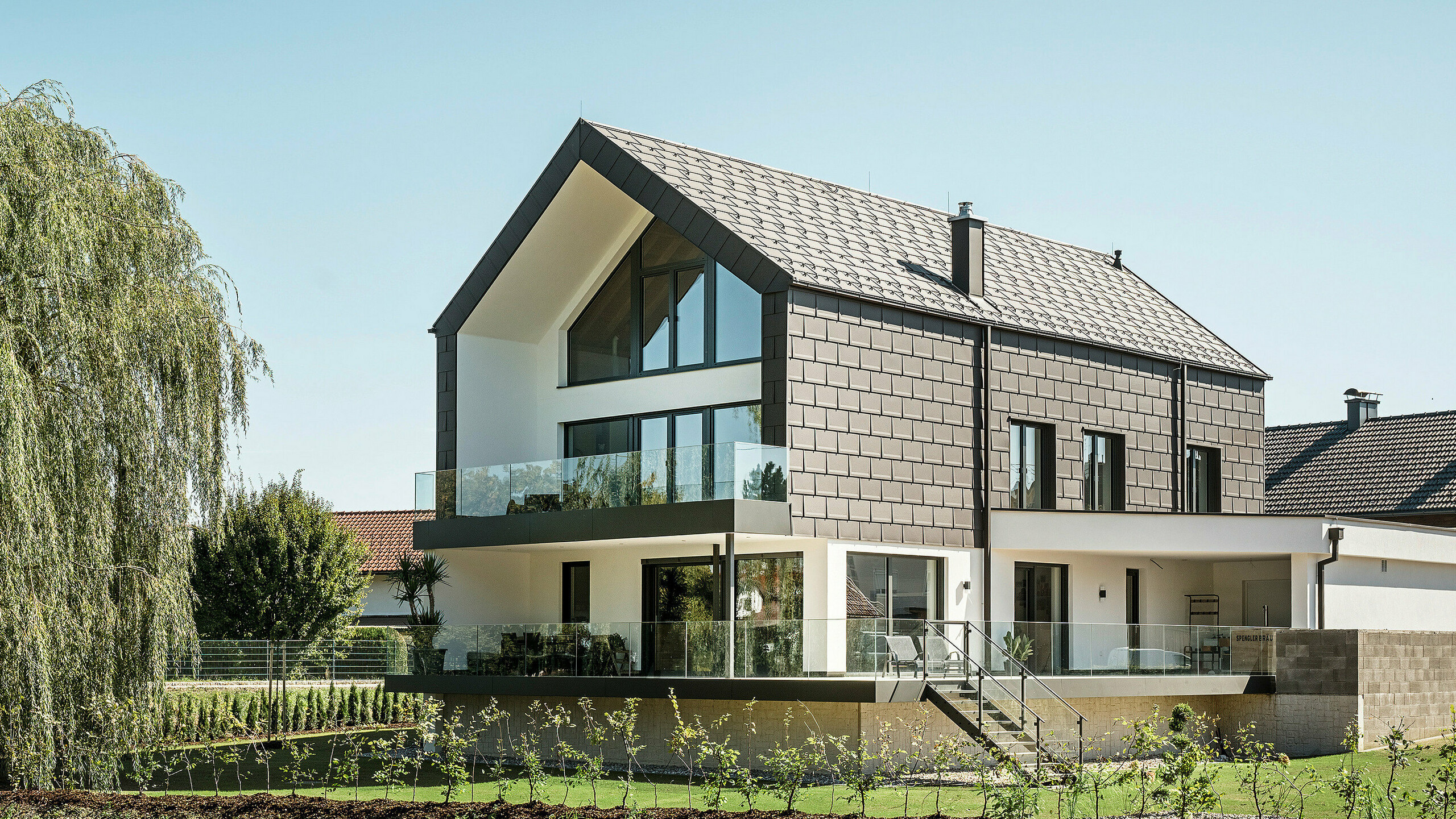 Einfamilienhaus in Neukirchen, Österreich, inmitten eines liebevoll angelegten Gartens. Die PREFA Dachplatte R.16 in braun P.10 fügt sich perfekt in das moderne Design des Hauses ein und schafft ein ästhetisches Zusammenspiel von traditionellen und modernen Stilelementen. Die großen Fenster öffnen das Gebäude zur umgebenden Natur und ermöglichen ein lichtdurchflutetes Wohnen. Die ruhige Wohnlage und die Einbindung der Gebäude in die natürliche Landschaft spiegeln die Ansprüche der Bauherren an die Lebensqualität wider.