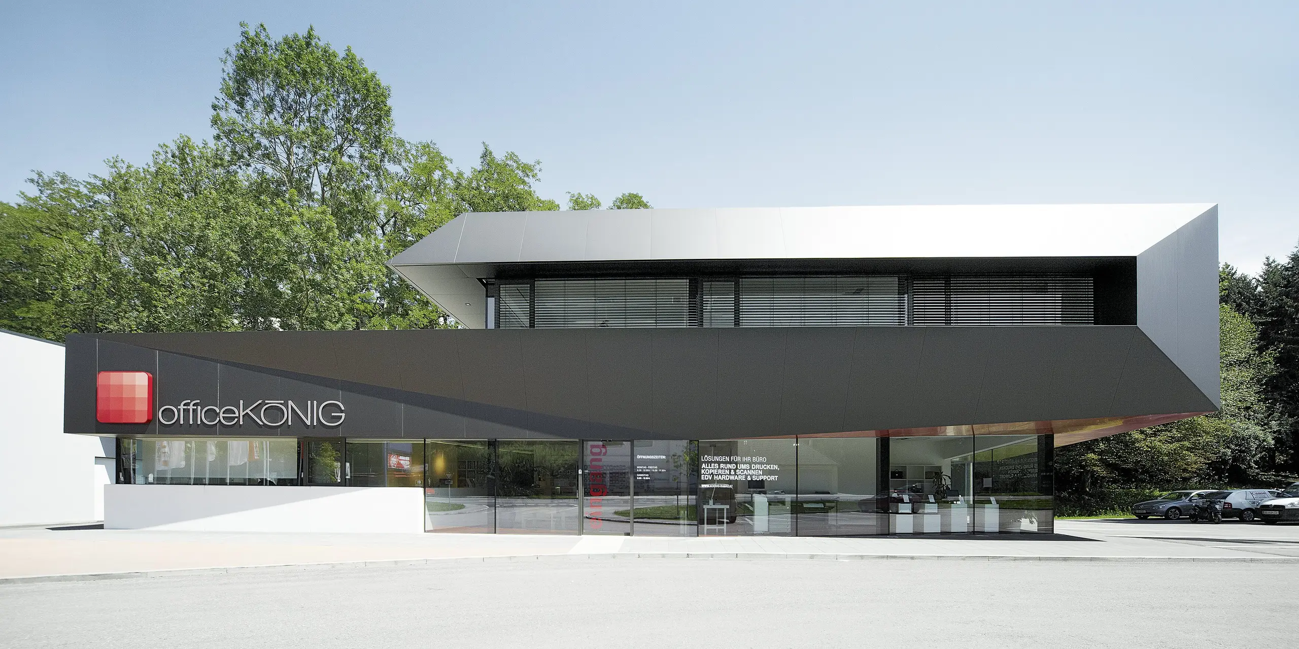 Modernes Firmengebäude von Office König in Bad Hall mit einer Fassade aus schwarzgrauen PREFA Aluminium Verbundplatten. Die langlebigen und witterungsbeständigen Platten verleihen dem Gebäude ein elegantes Aussehen und unterstreichen das innovative Design. Der Eingangsbereich wird durch eine großzügige Glasfront, das Firmenlogo und eine Terrasse über dem ersten Obergeschoss geprägt.