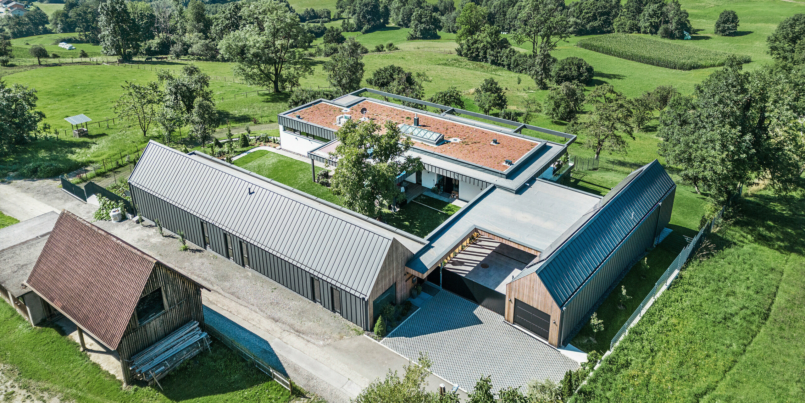 Letecký pohled na prostorný rodinný dům v kombinaci se zemědělskými halami v rakouském Pogier, zasazený do malebné krajiny s výhledem na zvlněné kopce a zelená pole. Budovy se vyznačují střešním a fasádním systémem PREFALZ v tmavě šedé barvě P.10, které vnášejí do venkovského prostředí moderní estetiku. Uspořádání budov vytváří příjemnou atmosféru dvora, zatímco FV panely na jedné ze střech odrážejí závazek domova k udržitelnosti a energetické účinnosti.