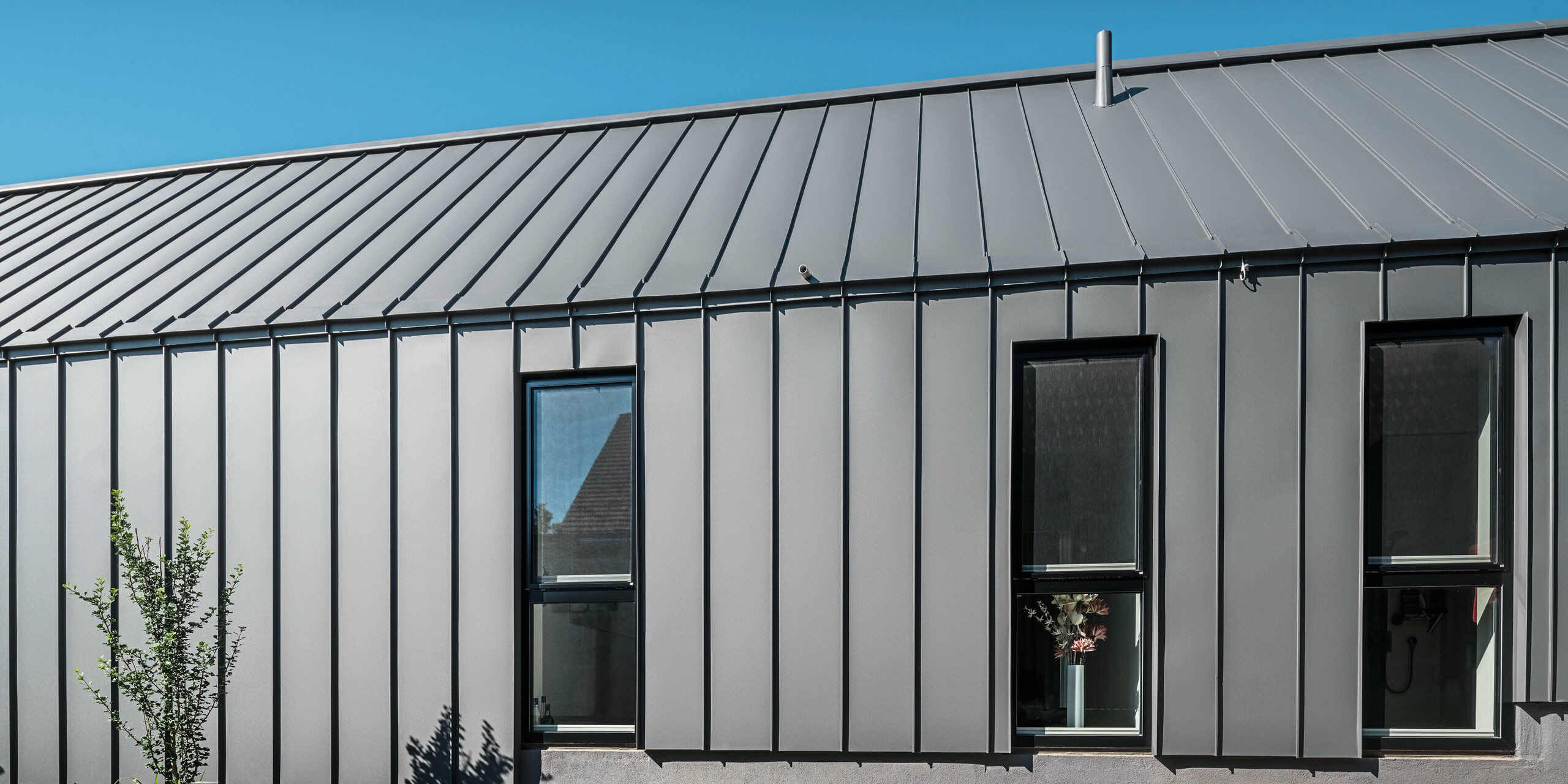 Boční pohled na rodinný dům v rakouském Pogier s odolným a stylovým PREFALZ v barvě P.10 tmavě šedá na střeše a fasádě. Hliníkový obklad se stojatou drážkou nabízí čisté vertikální linie, které elegantně rámují okna a vytvářejí moderní vzhled. Minimalistická architektura se nachází pod jasnou, jasně modrou oblohou.