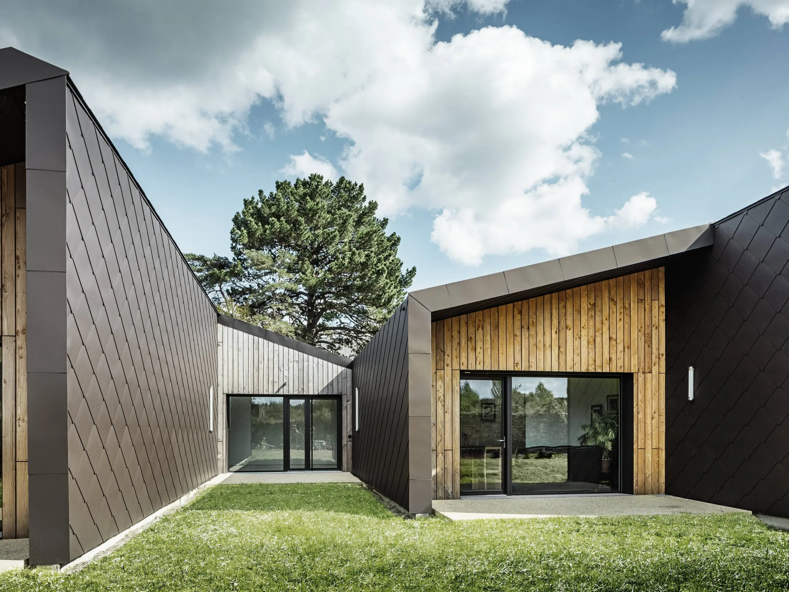 Das Bild zeigt moderne Architektur mit einer Fassade aus PREFA-Dach- und Wandelementen (44 × 44 mm) in Walnussbraun. Die Rautenstruktur der Aluminiumverkleidung verleiht dem Gebäude ein elegantes, geometrisches Erscheinungsbild. Die Fassade verbindet harmonisch natürliche Holzelemente mit großen Glasflächen und schafft so eine warme und einladende Atmosphäre. Der grüne Rasen und der klare Himmel unterstreichen den zeitgenössischen und zugleich naturnahen Charakter der Architektur.