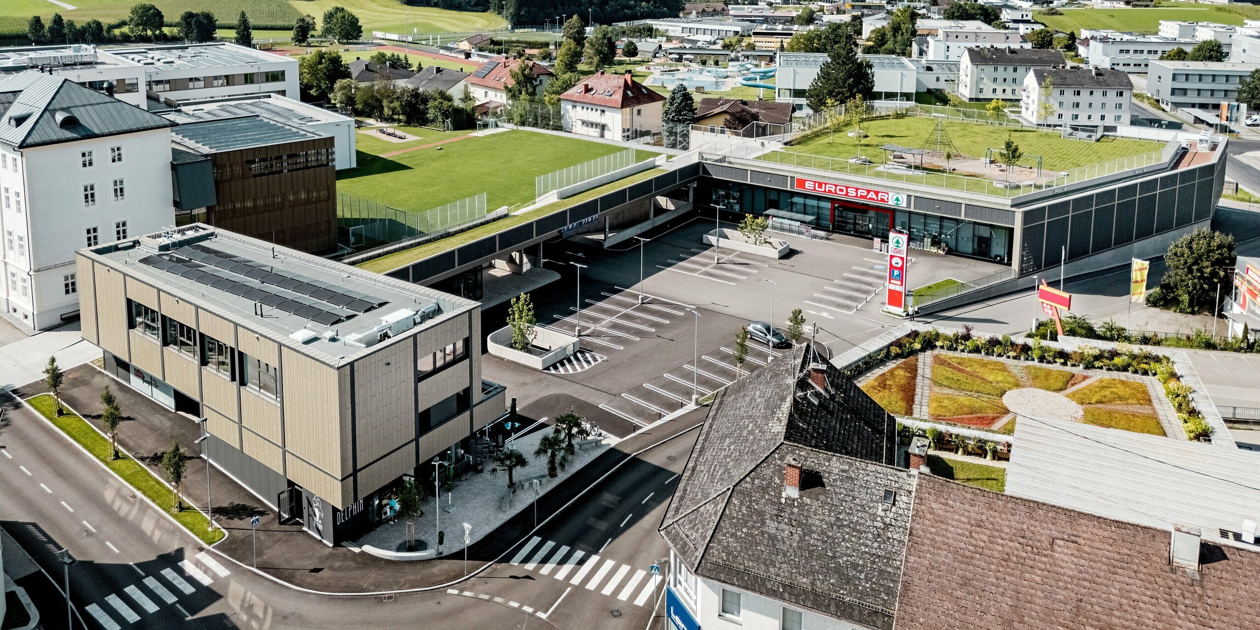 Die beeindruckende Luftaufnahme des Einkaufszentrums in Rohrbach verdeutlicht, wie es sich harmonisch in die ländliche Umgebung einfügt. Im Vordergrund ist der Gebäudekomplex mit seiner markanten Fassade aus PREFA Siding X-Paneelen in den Farben Anthrazit und Bronze zu sehen. Diese verleihen dem Gebäudeensemble eine moderne und elegante Ausstrahlung. Direkt davor erstreckt sich ein großzügiger Parkplatz, der von der klar strukturierten Architektur des Gebäudes eingerahmt wird. Der grüne Rasenbereich und die gepflegten Gartenanlagen rund um das Gebäude schaffen eine einladende Atmosphäre. Die umgebenden Häuser und das weite, hügelige Gelände im Hintergrund unterstreichen die gelungene Einbindung des modernen Bauwerks in seine Umgebung.