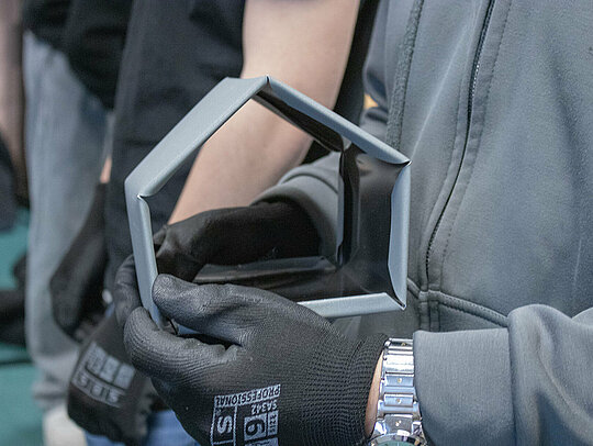 Ein Schüler hält ein fast fertiges Aluminium-Vogelhaus in den Händen, das er während der Handwerker-Tour 2025 gebaut hat. Das Aluminiumteil hat bereits die Form eines Hauses, aber es fehlen noch die letzten Schritte der Fertigung. Der Schüler trägt Schutzhandschuhe, und im Hintergrund sind weitere Teilnehmer der Handwerker-Tour zu erkennen.