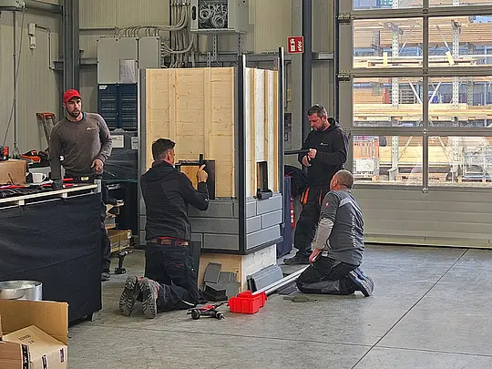 Vier Handwerker arbeiten in einer Werkhalle gemeinsam an einem aufgestellten PREFA Trainingsmodul. Zwei Personen knien am Boden und montieren Sidings an der unteren Konstruktion, während zwei weitere Fachkräfte im Stehen an den seitlichen Bauteilen arbeiten und Elemente ausrichten bzw. befestigen. Rund um das Modul liegen Werkzeuge wie Akkuschrauber, Blechprofile und eine rote Materialbox. Im Hintergrund sind große Hallentore.