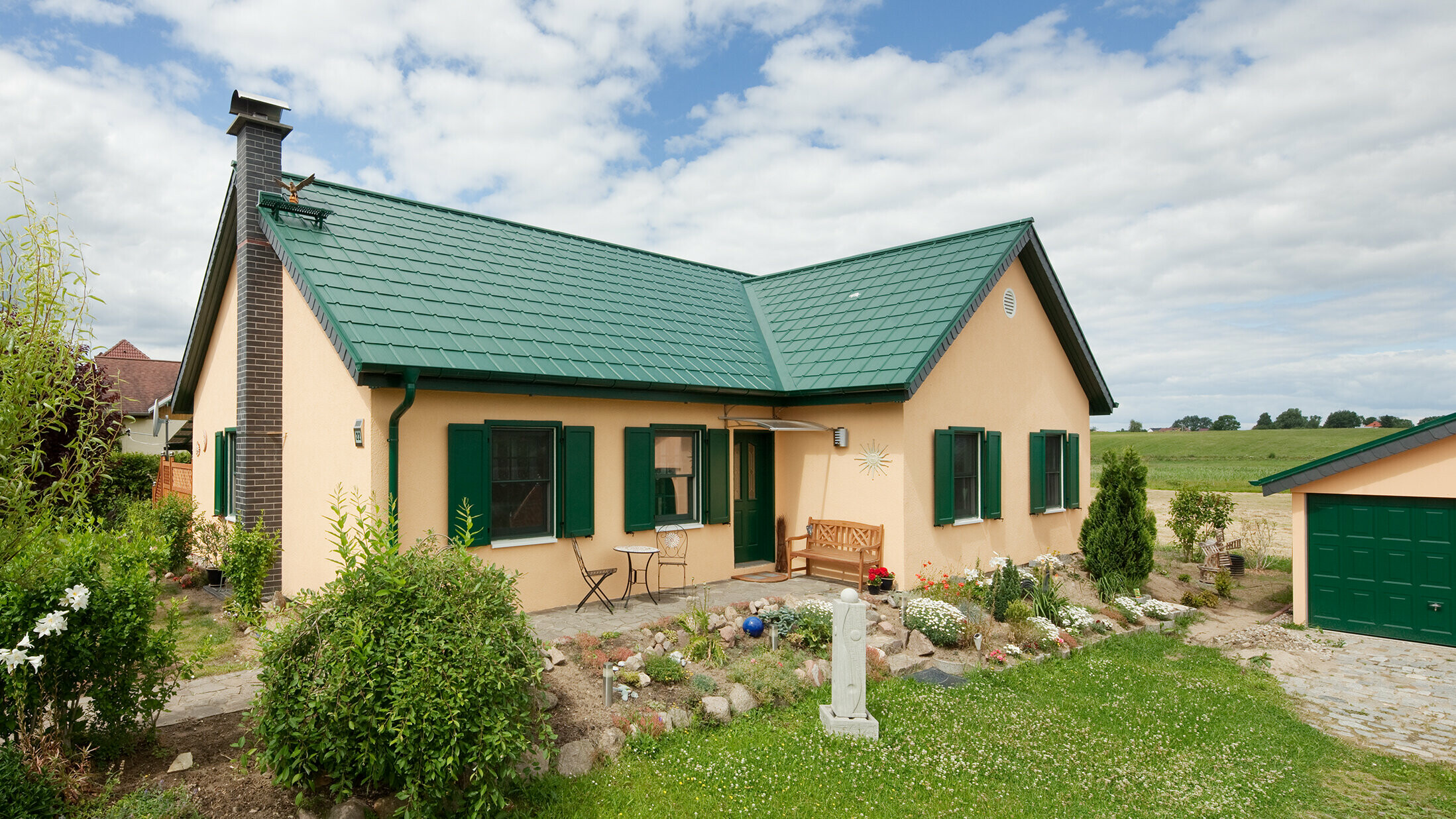 Die reizvolle Gesamtansicht des Einfamilienhauses in Friedrichswalde zeigt das langlebige Aluminiumdach in Moosgrün, das sowohl das Wohnhaus als auch die Garage ziert. Der grüne Farbton der PREFA Dachplatten fügt sich harmonisch in die natürliche Umgebung ein und unterstreicht die schlichte Architektur des Hauses. Das erweiterte Satteldach in L-Form liegt vor einem Feld. Das schmucke Häuschen hat passende grüne Fensterläden und einen hübschen kleinen Vorgarten.