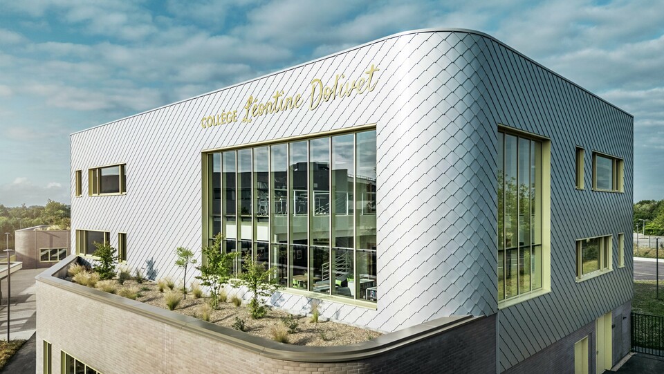 Das Foto zeigt eine markante Ecke des Collège Léontine Dolivet in Cesson-Sévigné. Die Fassade besteht aus silbermetallischen PREFA-Wandelementen, die diagonal angeordnet sind und das Licht reflektieren. Besonders auffällig ist die geschwungene Form des Obergeschosses, die dem Gebäude eine sanfte und moderne Anmutung verleiht. Große Glasflächen geben den Blick ins Innere frei, während gelbe Fensterrahmen farbliche Akzente setzen. Im Vordergrund befindet sich eine bepflanzte Fläche mit Kies und Sträuchern, die die Architektur harmonisch ergänzt. Der leicht bewölkte Himmel hebt die Metalloberfläche zusätzlich hervor.