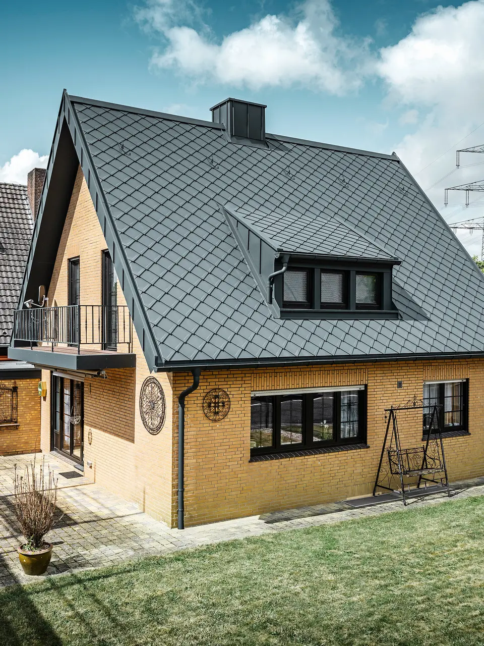 Ein Einfamilienhaus in Tönisvorst mit einem PREFA Dach aus Dachrauten 29×29 in P.10 Anthrazit, das ein herausstechendes Rautenmuster bietet. Schleppgauben, Kamin und der Eingangsbereich sind mit PREFALZ verkleidet, was zusammen mit Ablaufrohr und Hängerinne in P.10 Anthrazit dem Haus einen zeitlosen und eleganten Look verleiht. Das Backsteingebäude mit dem Aluminiumdach harmoniert mit der ruhigen, grünen Umgebung der Siedlung.