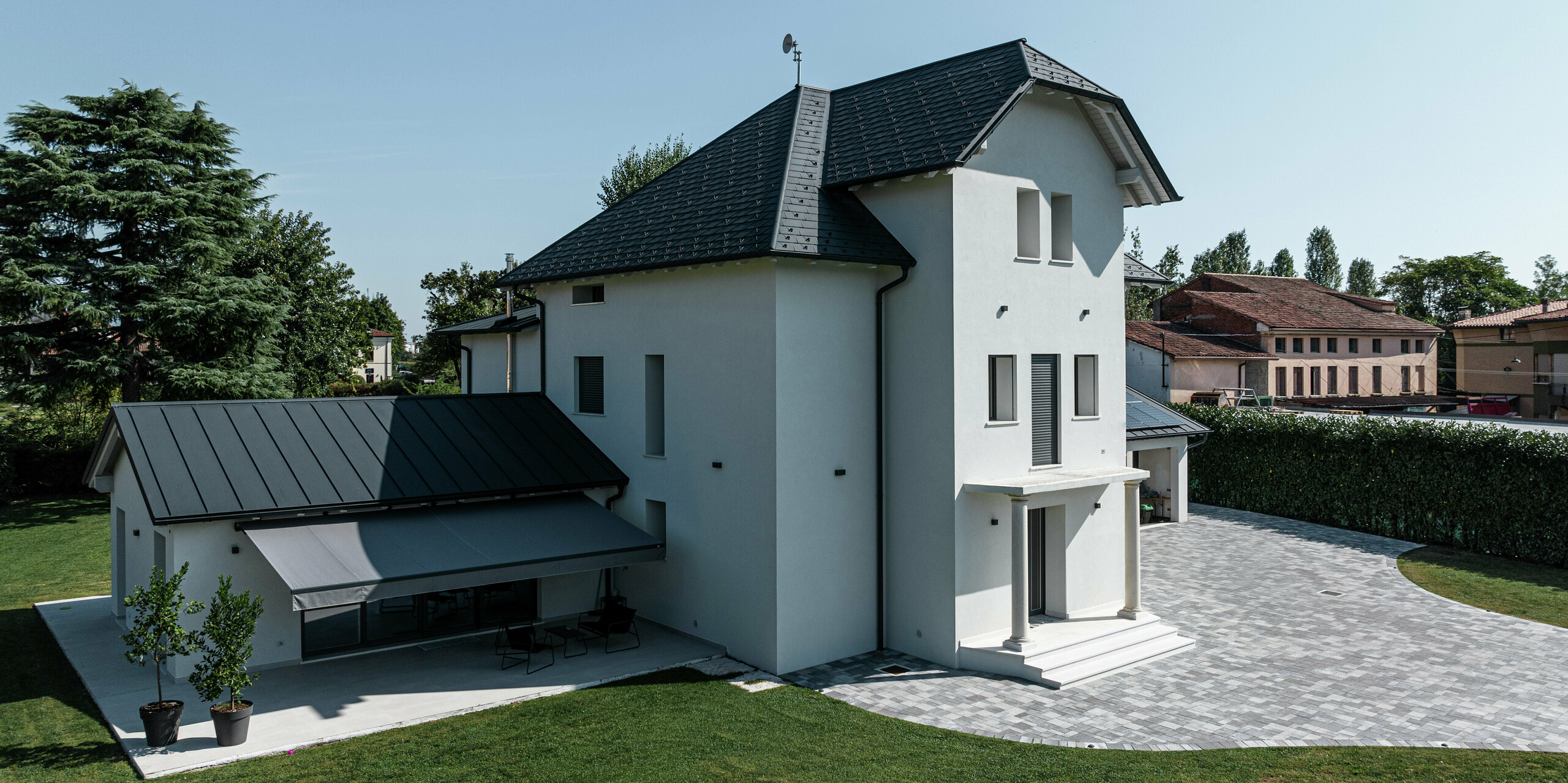 Seitenansicht eines modernen Einfamilienhauses in Marano Vicentino, Italien, mit robusten PREFA Dachschindeln in P.10 Anthrazit. Das angrenzende Nebengebäude wurde mit einer langlebigen Doppelstehfalzdeckung ausgestattet. Die hellgraue Markise spendet Schatten bei starker Mittagssonne. Die klare Architektur und die hochwertigen Aluminiumprodukte sorgen für eine moderne und witterungsbeständige Dachlandschaft.