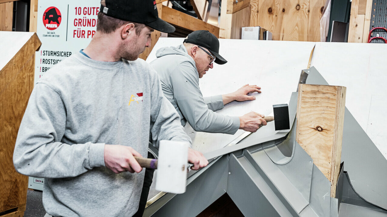 Zwei Teilnehmer des PREFA Academy Kurses arbeiten an einem Modell eines Daches. Sie verwenden Werkzeuge wie Gummihämmer, um Dachelemente aus Aluminium präzise anzubringen. Im Hintergrund ist ein Schild mit den Vorteilen von PREFA-Produkten zu sehen. Die Szene zeigt die praktische Anwendung und den handwerklichen Unterricht in der Akademie.