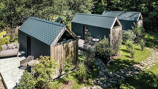 Luftaufnahme mehrerer minimalistischer Tiny Houses im Secret Koi Garden am Natterer See, eingebettet in dichtes Grün und naturnahe Gartenwege. Die Dach‑ und Fassadenflächen sind vollständig mit PREFA Dach‑ und Wandrauten aus Aluminium in P.10 Anthrazit verkleidet und prägen mit ihrer gleichmäßigen, fischhautartigen Struktur das Erscheinungsbild. Das leichte, langlebige und witterungsbeständige PREFA Aluminium ermöglicht präzise Details und unterstreicht die klare Glamping‑Architektur im Einklang mit der Natur.