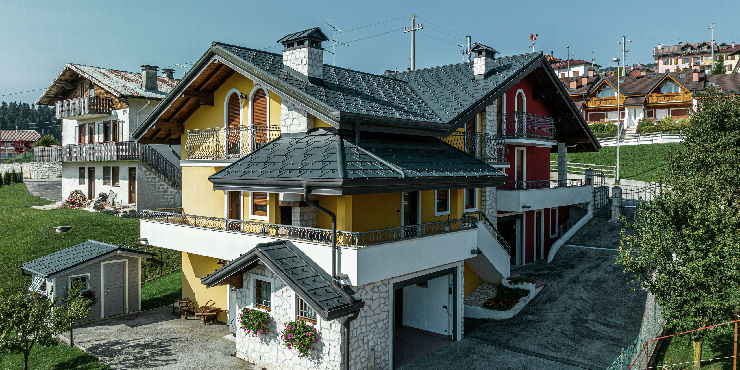Einfamilienhaus in Gallio im alpinen Architekturstil mit modernem PREFA Blechdach in P.10 Dunkelgrau. PREFA Dachplatten R.16 sorgen für eine langlebige, witterungsbeständige und optisch ansprechende Dachlösung. Die harmonische Kombination aus gelb-roter Fassade, Natursteinelementen und Holzakzenten verleiht dem Haus einen einladenden Charakter. Die PREFA Dachentwässerung in P.10 dunkelgrau sorgt für eine zuverlässige Wasserableitung. Ein Paradebeispiel für ästhetische und funktionale Architektur in alpiner Umgebung.
