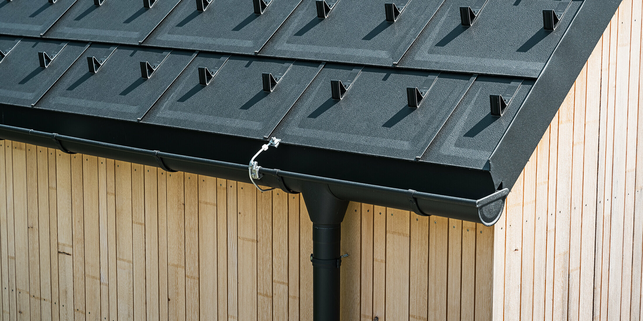 Nahaufnahme der PREFA Dachplatte R.16 mit Schneestoppern, Dachrinne und Ablaufrohr in Schwarz P.10 inkl. Blitzschutz und einer senkrechten Holzfassade. Die robusten PREFA Aluminiumprodukte schmücken das Satteldach eines Einfamilienhauses in Lustenau, Vorarlberg.