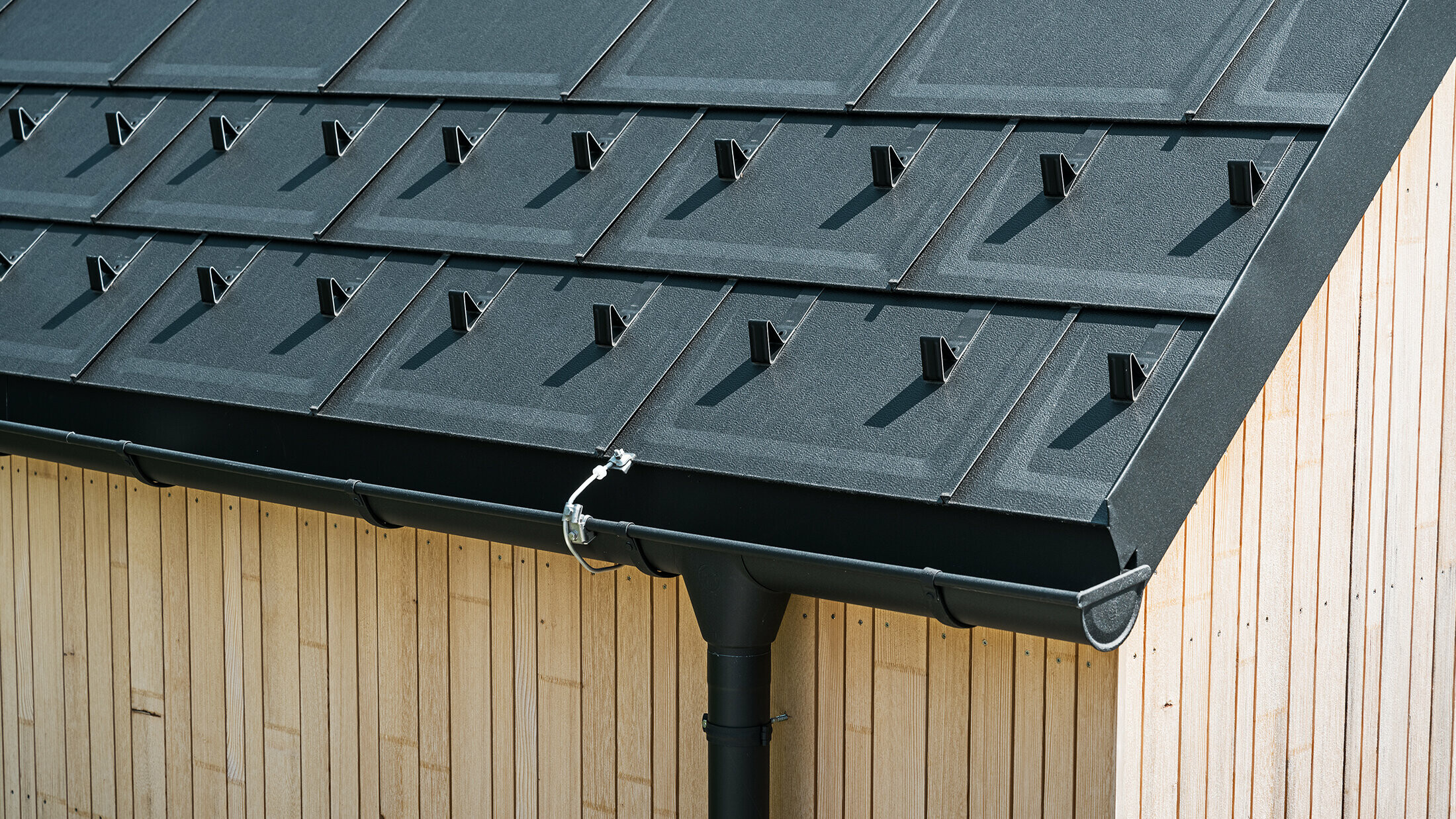 Nahaufnahme der PREFA Dachplatte R.16 mit Schneestoppern, Dachrinne und Ablaufrohr in Schwarz P.10 inkl. Blitzschutz und einer senkrechten Holzfassade. Die robusten PREFA Aluminiumprodukte schmücken das Satteldach eines Einfamilienhauses in Lustenau, Vorarlberg.