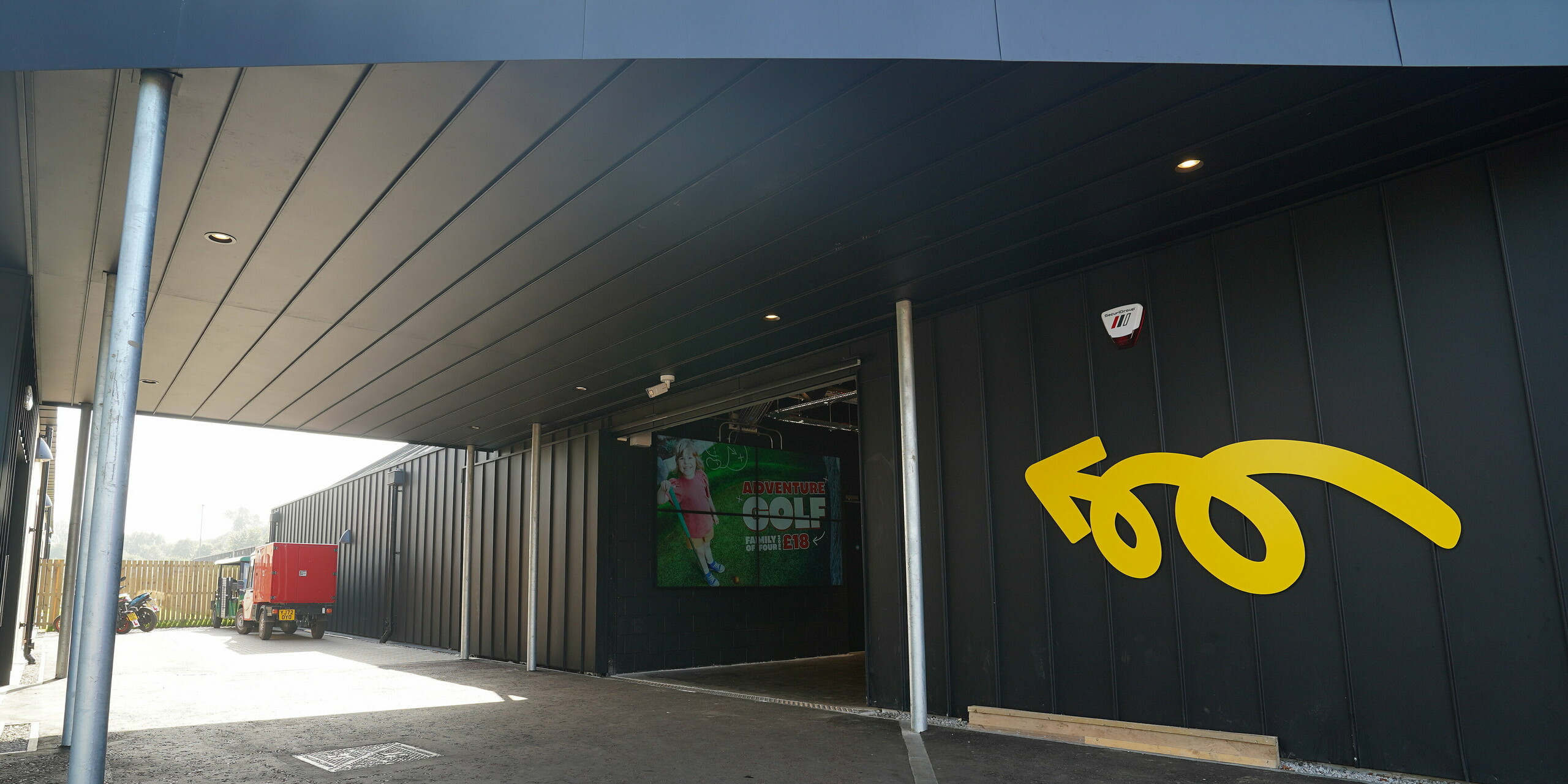 Eingangsbereich der 'Golf it!' Golfanlage in Glasgow, Schottland, mit einem modern gestalteten Vordach aus PREFALZ in P.10 Schwarz. Die Aluminium-Elemente schaffen eine einladende Atmosphäre, die durch einen geschwungenen Pfeil in Gelb an der Wand und ein großflächiges Eingangsschild akzentuiert wird. Diese Details zeigen, wie PREFALZ ästhetisch und funktionell in unterschiedlichen Gebäudebereichen integriert werden kann.