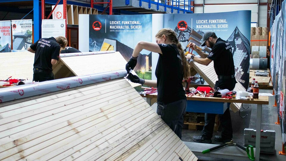 Teilnehmende der Deutschen Meisterschaft im Klempnerhandwerk arbeiten in der PREFA Academy in Wasungen an Übungsdächern aus Holz und Aluminium. Auf den Arbeitstischen liegen Werkzeuge und Materialien bereit. Im Hintergrund sind PREFA-Banner zu sehen.