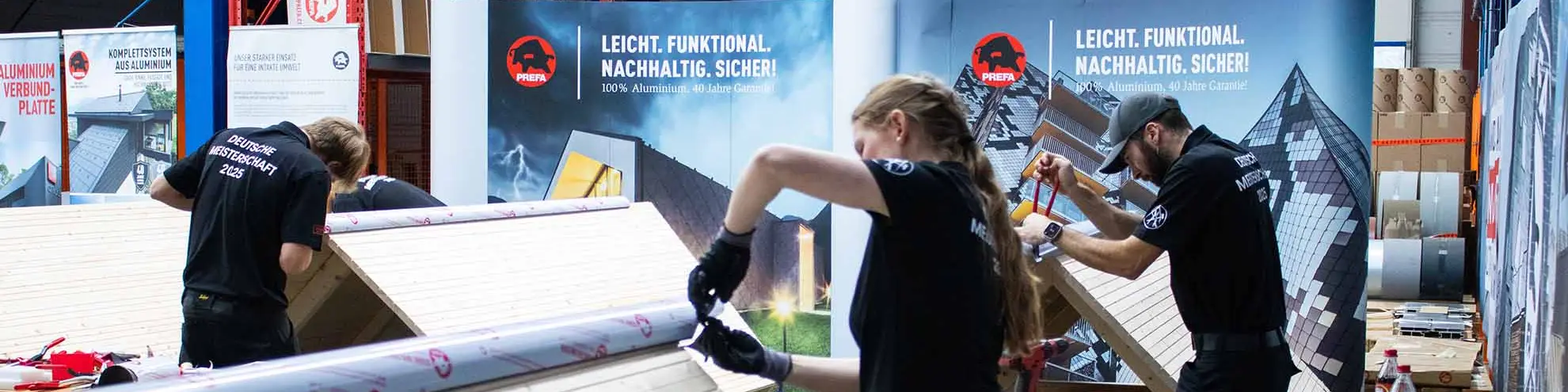 Teilnehmende der Deutschen Meisterschaft im Klempnerhandwerk arbeiten in der PREFA Academy in Wasungen an Übungsdächern aus Holz und Aluminium. Auf den Arbeitstischen liegen Werkzeuge und Materialien bereit. Im Hintergrund sind PREFA-Banner zu sehen.