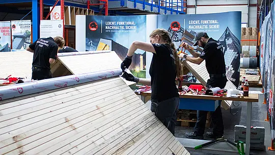 Teilnehmende der Deutschen Meisterschaft im Klempnerhandwerk arbeiten in der PREFA Academy in Wasungen an Übungsdächern aus Holz und Aluminium. Auf den Arbeitstischen liegen Werkzeuge und Materialien bereit. Im Hintergrund sind PREFA-Banner zu sehen.