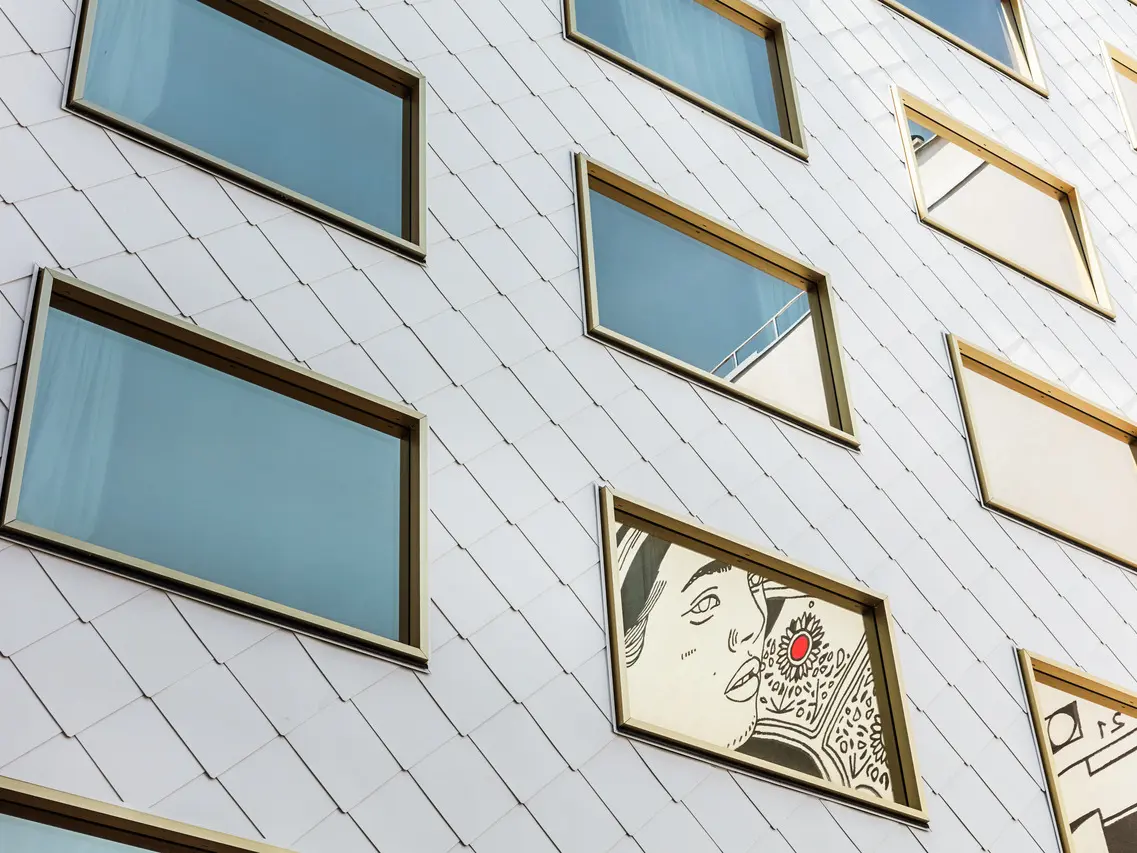 Detailaufnahme der Fassade des "THE ROCK Radisson RED Vienna" Hotels, das mit PREFA Dach- und Wandrauten 44 × 44 in P.10 Reinweiß verkleidet ist. Unter den mehreren spiegelverglasten Fenstern mit goldenen Rahmen sticht ein Fenster hervor, in das ein Kunstwerk eingelassen ist, welches das Porträt einer Person mit einem markanten roten Akzent darstellt