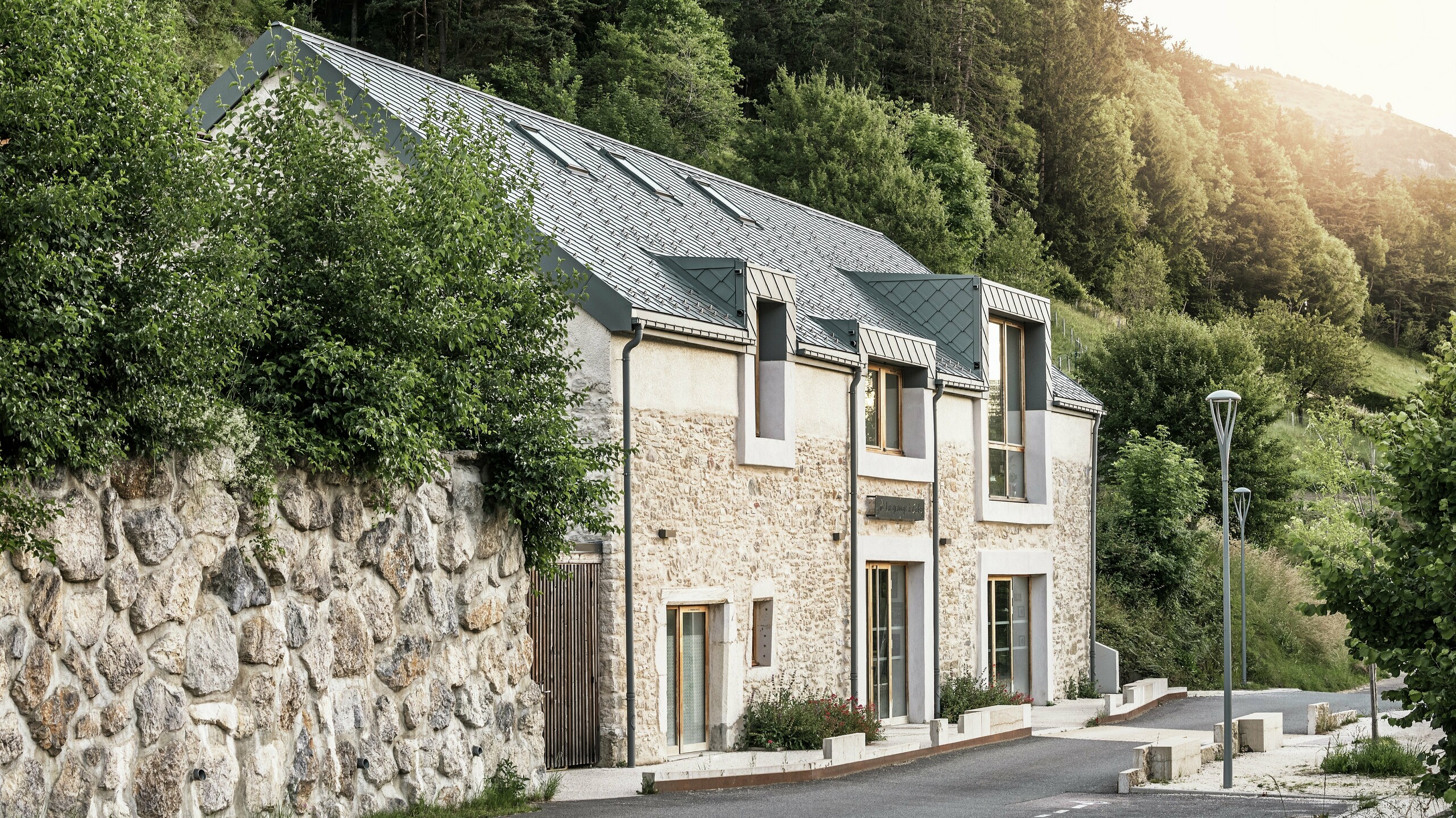 Seitenansicht der sanierten Scheune "La Grange à Gaby" in Engins, Frankreich. Das Gebäude kombiniert traditionelles Steinemauerwerk mit einem modernen PREFA Aluminiumdach aus 29 × 29 cm großen Rauten. Die behutsame Renovierung verbindet historische Bauweise mit langlebigen Materialien.