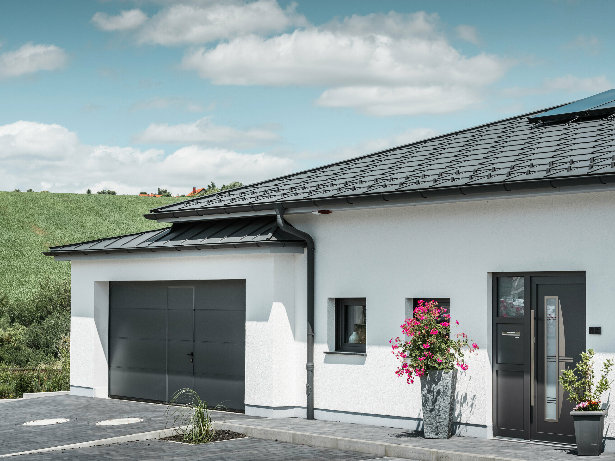 Das Bild präsentiert die Vorderseite eines Einfamilienhauses in Ilmenau, Thüringen, mit einem eleganten PREFA Dach in P.10 Dunkelgrau. Die Dachplatte R.16 verleiht dem Haus ein modernes Aussehen, während das integrierte PREFA Dachentwässerungssystem mit seiner dunkelgrauen Hängerinne und dem Ablaufrohr effizienten Abfluss von Regenwasser garantieren. Die Garage, ausgestattet mit dunkelgrauem PREFALZ, fügt sich harmonisch in die Gesamtästhetik ein und unterstreicht die Qualität und den Stil von PREFA Aluminiumprodukten.