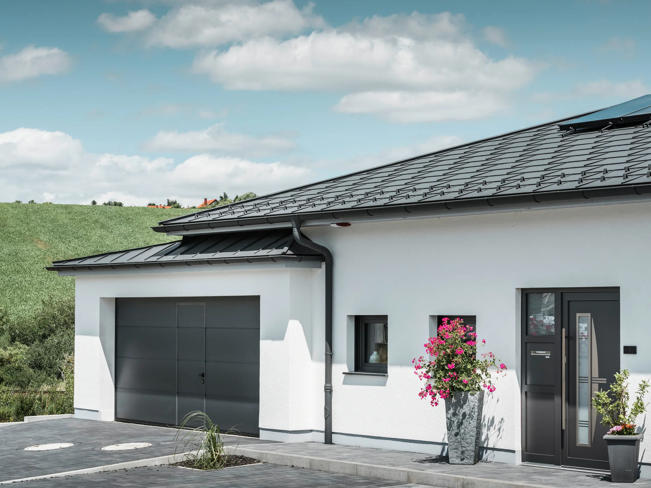 Das Bild präsentiert die Vorderseite eines Einfamilienhauses in Ilmenau, Thüringen, mit einem eleganten PREFA Dach in P.10 Dunkelgrau. Die Dachplatte R.16 verleiht dem Haus ein modernes Aussehen, während das integrierte PREFA Dachentwässerungssystem mit seiner dunkelgrauen Hängerinne und dem Ablaufrohr effizienten Abfluss von Regenwasser garantieren. Die Garage, ausgestattet mit dunkelgrauem PREFALZ, fügt sich harmonisch in die Gesamtästhetik ein und unterstreicht die Qualität und den Stil von PREFA Aluminiumprodukten.