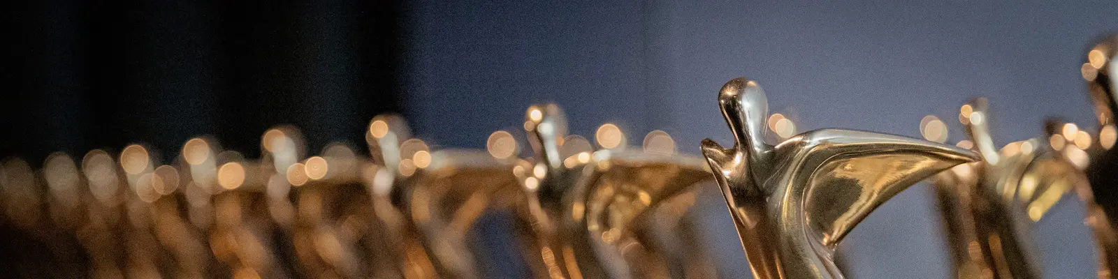 Das Bild zeigt eine Reihe von Trophäen aus Gold, welche stilisierte menschliche Figuren darstellen, die eine triumphierende Pose einnehmen. 