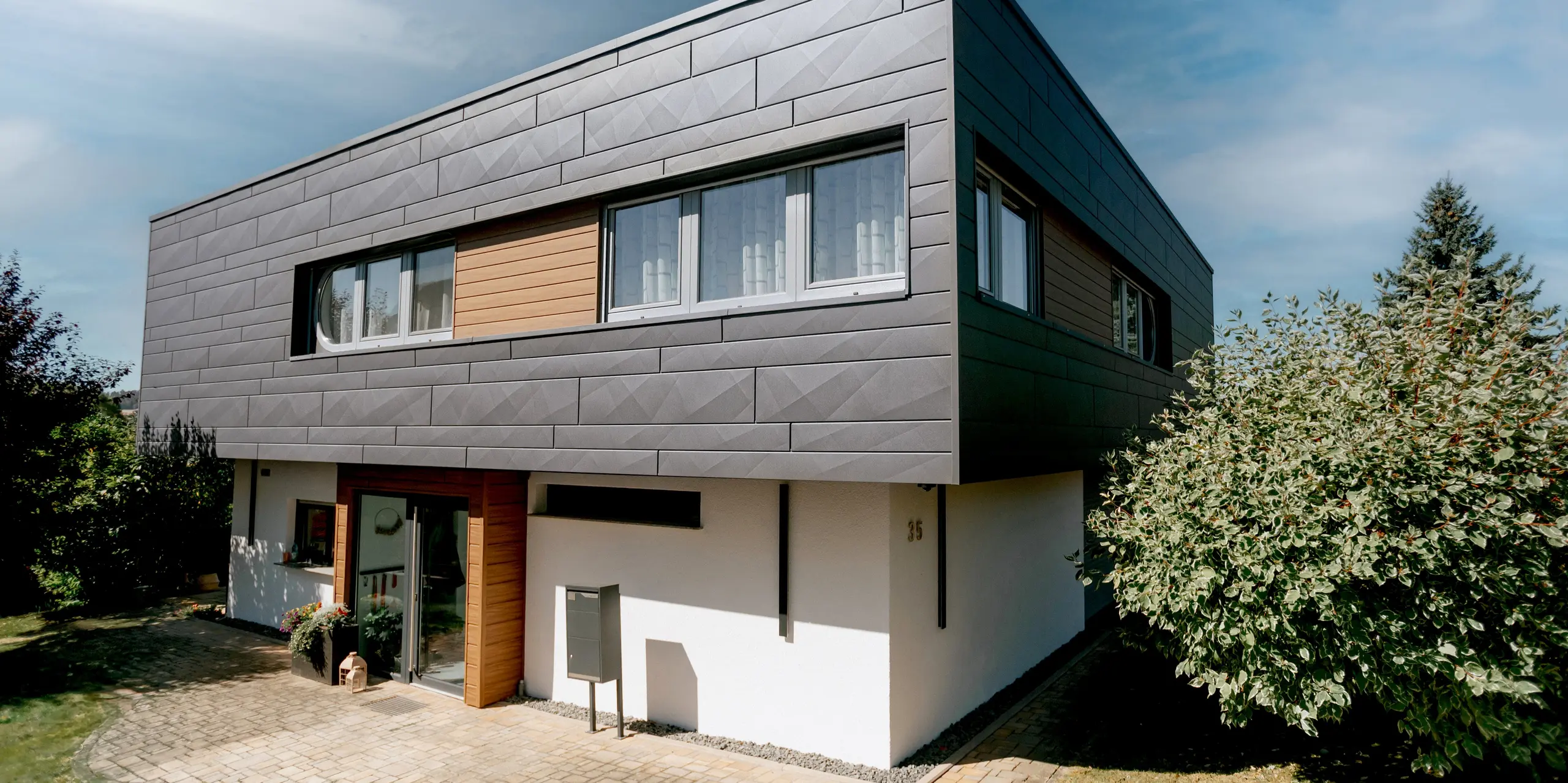 Modernes Einfamilienhaus in Marksuhl mit ausdrucksstarker PREFA Aluminiumfassade: Die horizontalen PREFA Siding.X Paneele in P.10 Dunkelgrau verleihen dem auskragenden Obergeschoss eine klare, elegante Struktur. Kombiniert mit Siding-Elementen in warmen Walnuss braun entsteht ein harmonisches Fassadenbild. Die langlebige, witterungsbeständige Aluminiumverkleidung schützt dauerhaft und überzeugt durch hochwertige Optik.