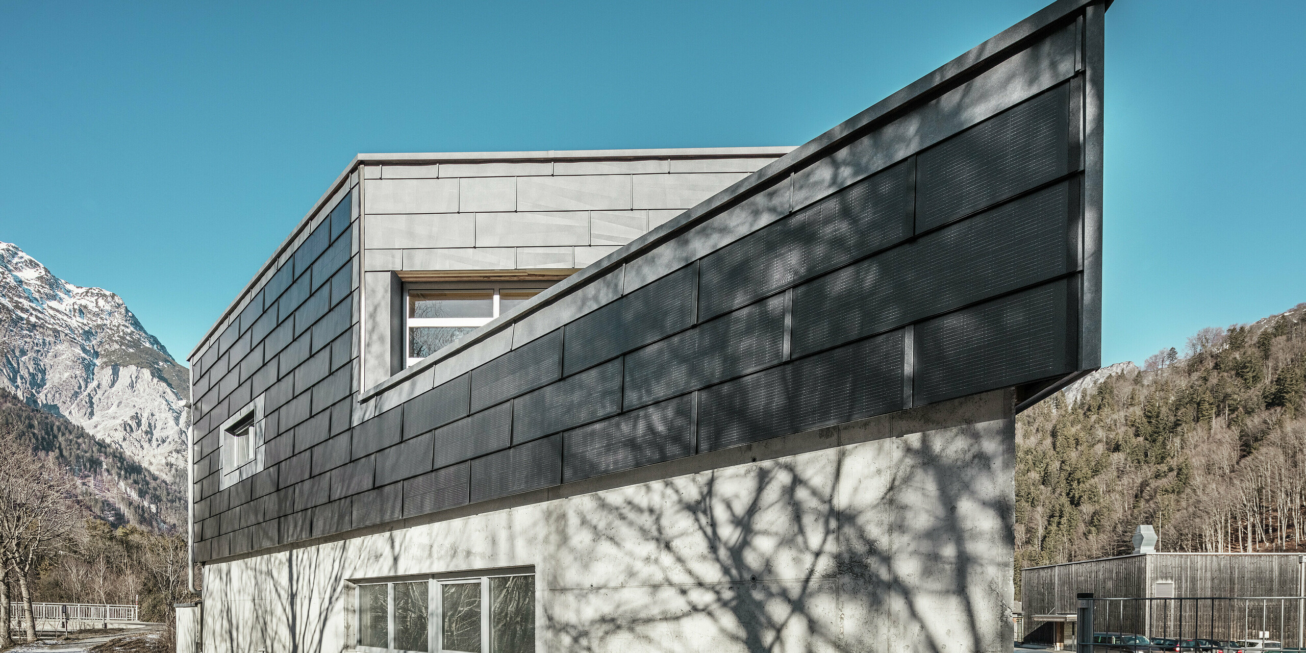 Das Firmengebäude der Spenglerei RÜMA in Vorarlberg beeindruckt mit seiner modernen Fassade aus PREFA Solardachplatten und Sichtbeton. Umgeben von Bergen und Bäumen, deren Schatten sich auf der Wand abzeichnen, steht es unter einem klaren, blauen Himmel. Die spitze Gebäudeecke und die PREFA Fassadenpaneele FX.12 betonen das markante Erscheinungsbild zusätzlich.