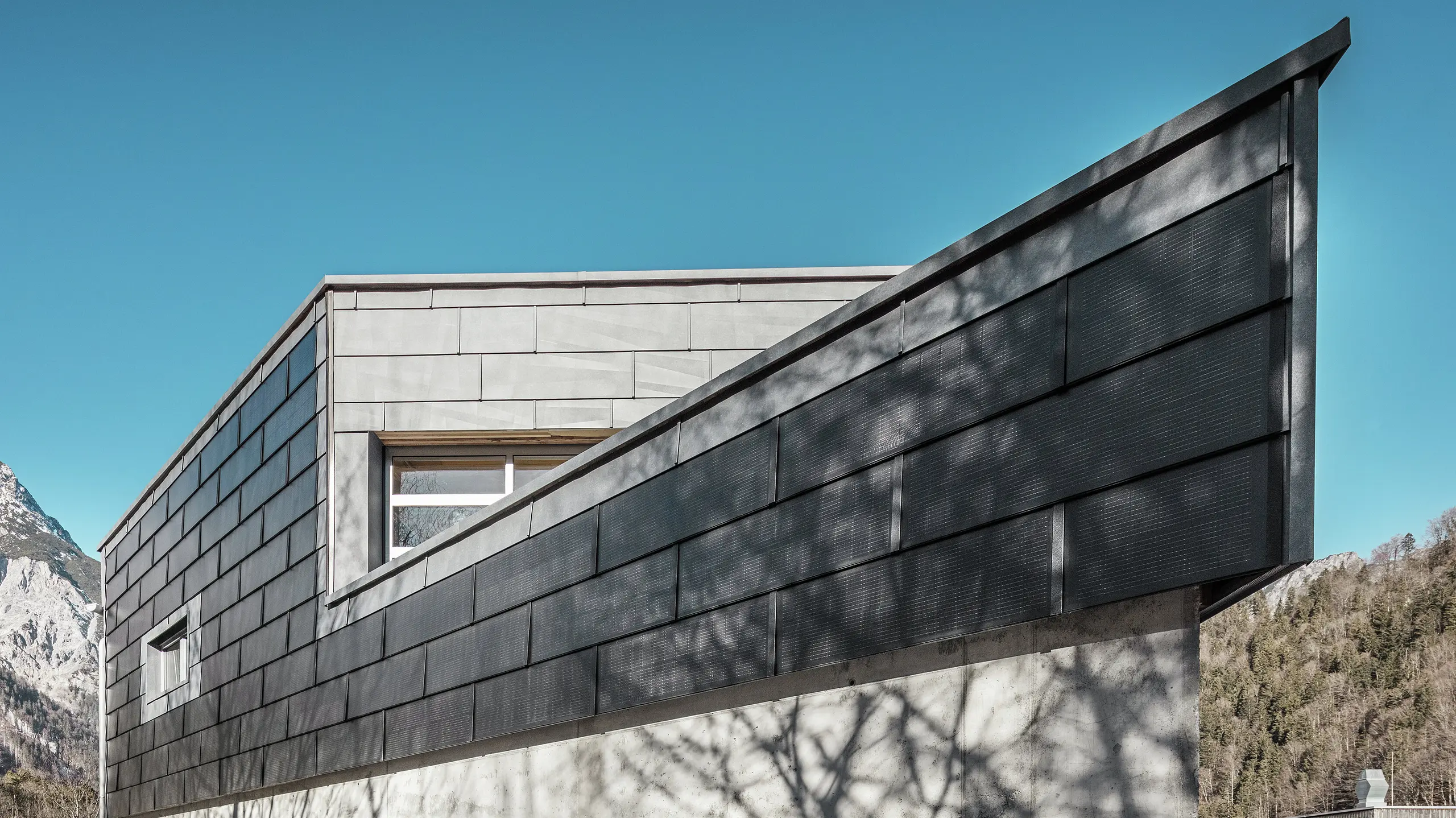 Das Bürogebäude der RÜMA Blechwerkstatt in Vorarlberg besticht durch seine moderne Fassade aus PREFA Solardachpaneelen und Sichtbeton. Umgeben von Bergen und Bäumen, deren Schatten sich an der Wand abzeichnen, erhebt es sich unter strahlend blauem Himmel. Die markante Gebäudekante und die PREFA Fassadenpaneele FX.12 unterstreichen den eindrucksvollen Eindruck zusätzlich.