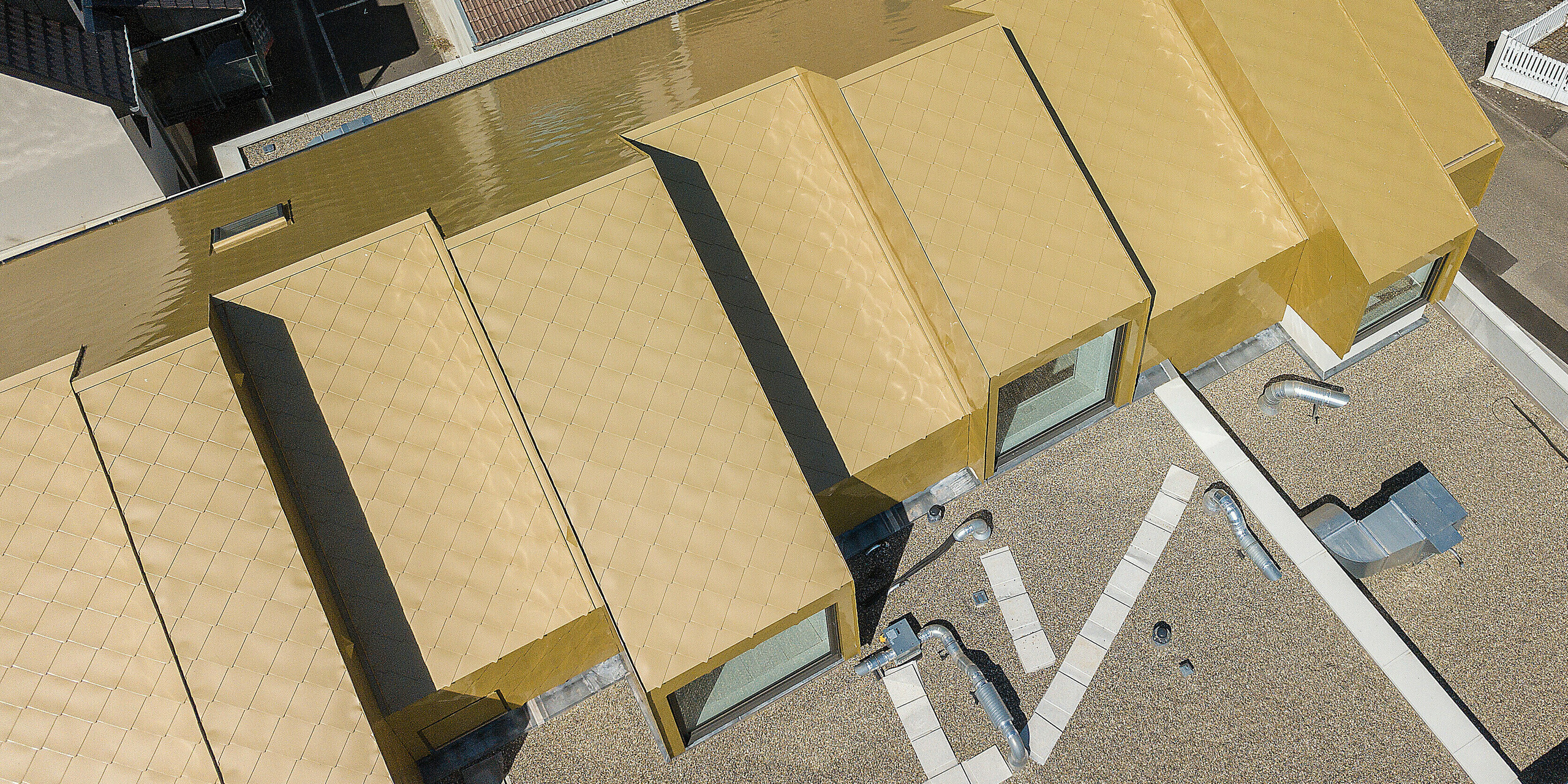 Blick von oben auf das goldene Dach des Kulturzentrums Fessenheim. Das neue Dach wurde mit PREFA Dachrauten 44 × 44 in Sparkling Gold verkleidet. Im Hintergrund sind benachbarte Gebäudedächer zu erkennen.