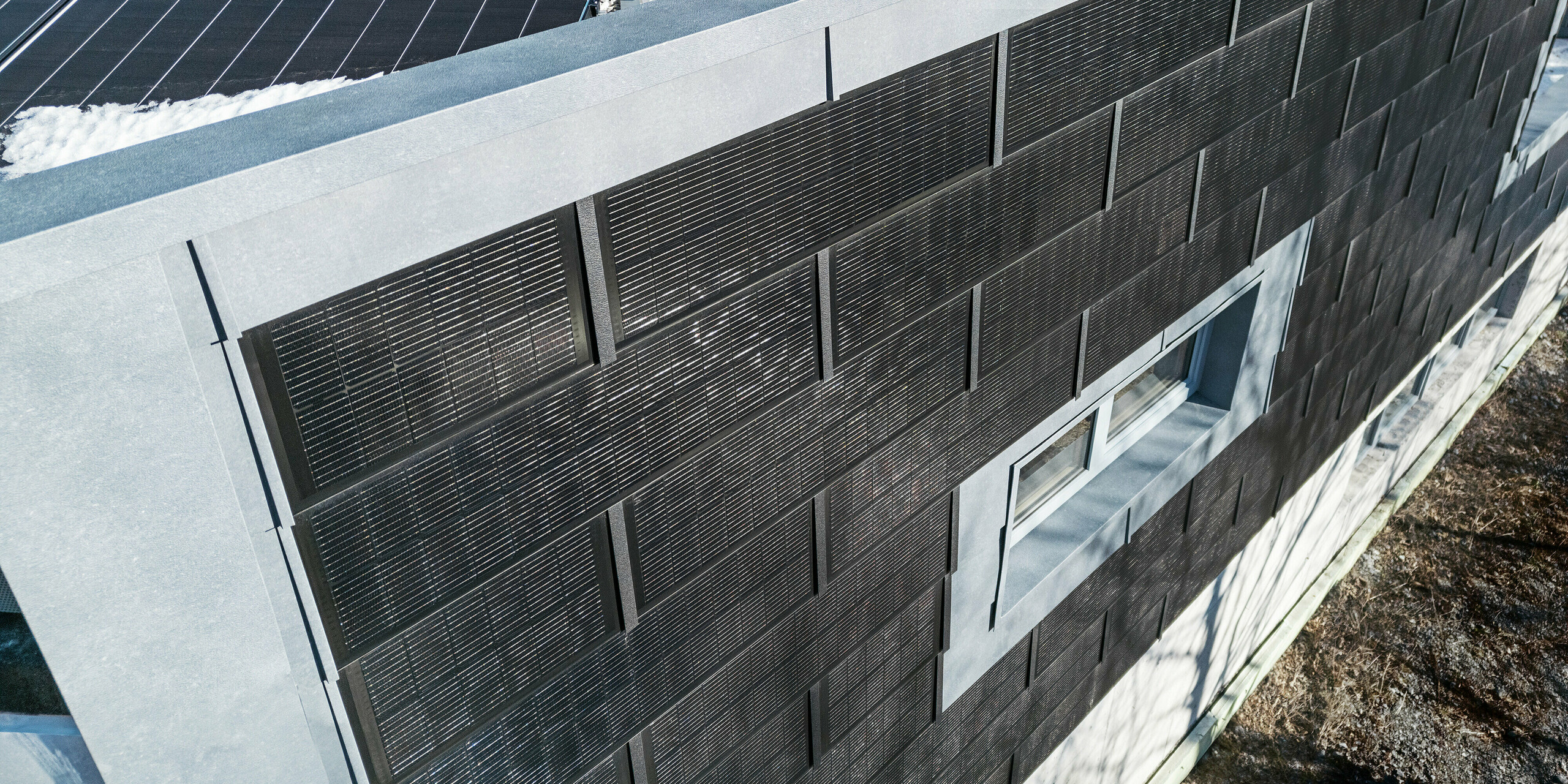 Die Draufsicht zeigt die Fassade der Spenglerei RÜMA in Vorarlberg mit grauen Fassadenpaneelen FX.12 sowie innovativen PREFA Solardachplatten mit integrierten Photovoltaikmodulen. Im Vordergrund ist das teilweise mit Schnee bedeckte Flachdach mit weiteren Photovoltaikmodulen zu sehen.