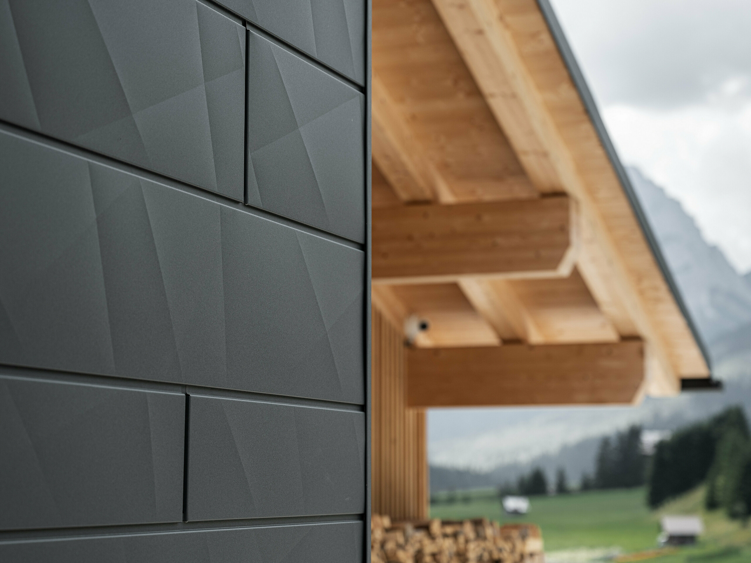 Nahaufnahme der Siding.X Fassadenpaneele in P.10 Schwarz von PREFA an der Casa Hoffe in Sappada, Italien. Das Fassadensystem aus Aluminium ist gekonnt in einem modernen, abstrakten Design angebracht, das einen ansprechenden Kontrast zu den sichtbaren Holzelementen des Dachstuhls bildet. Die innovative Fassadenlösung fügt sich nahtlos in das alpine Ambiente ein und steht für die Verbindung von Funktionalität, Beständigkeit und ästhetischer Baukultur.