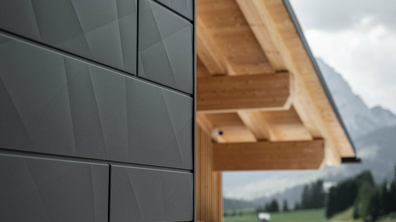 Nahaufnahme der Siding.X Fassadenpaneele in P.10 Schwarz von PREFA an der Casa Hoffe in Sappada, Italien. Das Fassadensystem aus Aluminium ist gekonnt in einem modernen, abstrakten Design angebracht, das einen ansprechenden Kontrast zu den sichtbaren Holzelementen des Dachstuhls bildet. Die innovative Fassadenlösung fügt sich nahtlos in das alpine Ambiente ein und steht für die Verbindung von Funktionalität, Beständigkeit und ästhetischer Baukultur.