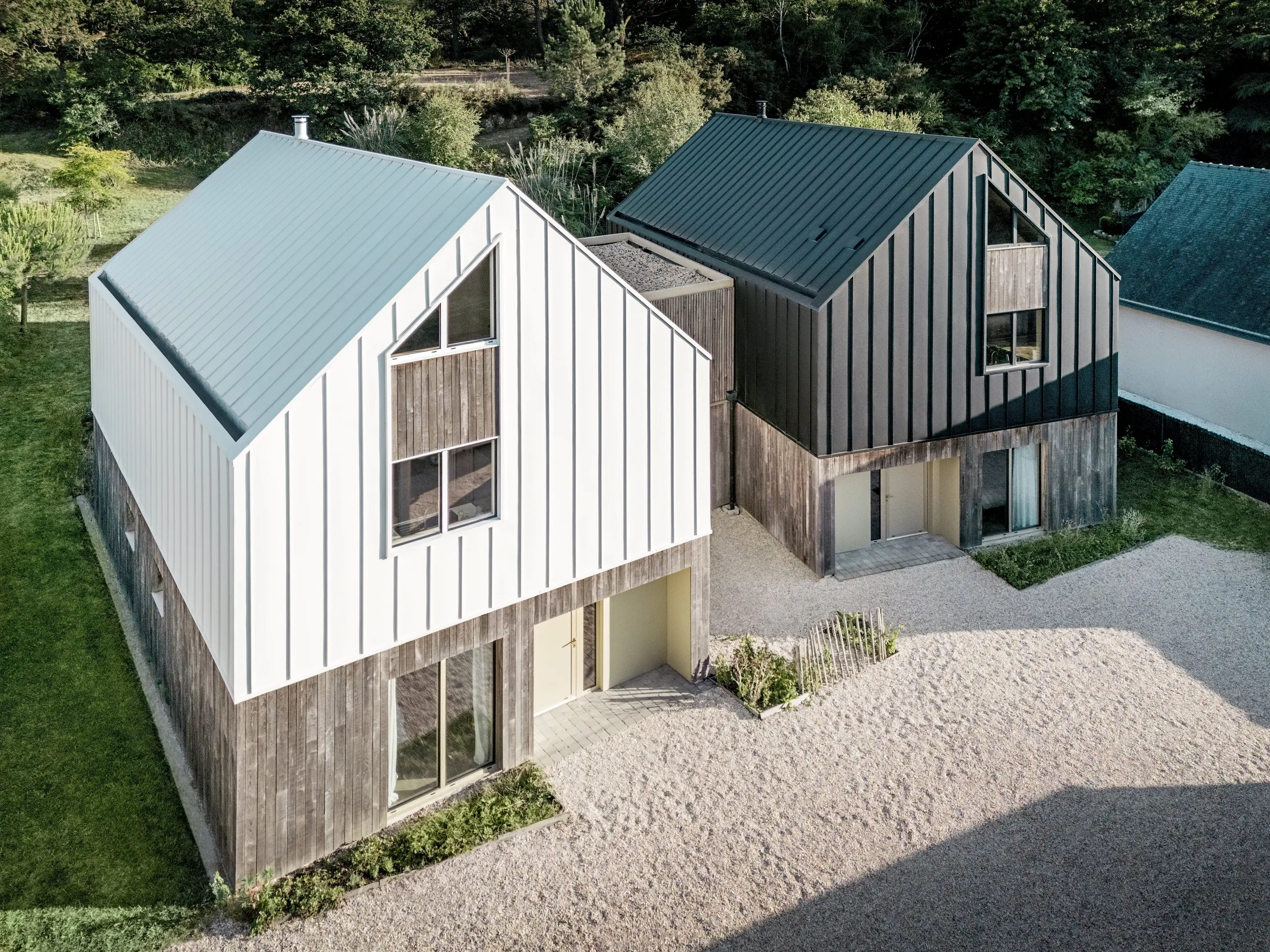 Die Luftaufnahme des Wohnensembles Le Rhune zeigt zwei moderne Häuser mit markanten Satteldächern und kontrastierenden PREFALZ Aluminiumhüllen in P.10 Reinweiß und Schwarzgrau. Die klaren Baukörper orientieren sich an traditionellen bretonischen Fischerhäusern und fügen sich harmonisch in die umgebende Landschaft ein. Besonders ins Auge fallen die durchgehenden Stehfalzlinien, die Dach und Fassade nahtlos miteinander verbinden und die präzise Geometrie betonen. Das langlebige, wetterbeständige PREFALZ Aluminium ermöglicht eine hochwertige Gebäudehülle mit klarer Formsprache, hoher Gestaltungsfreiheit und dauerhaft ansprechender Optik.