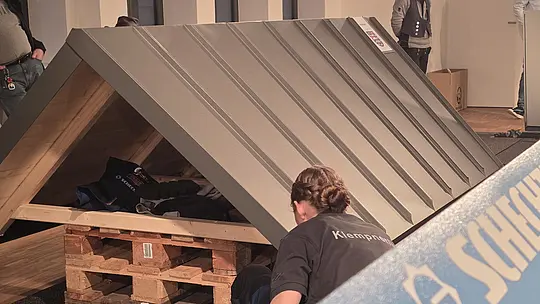 Eine Teilnehmerin arbeitet an einem Dachmodell, das auf einer Holzpalette steht. Das Blechdach ist bereits verlegt, und die Person überprüft die letzte Details, um eine perfekte Ausführung zu erreichen. Im Hintergrund sind andere Teilnehmer bei der Arbeit zu sehen.