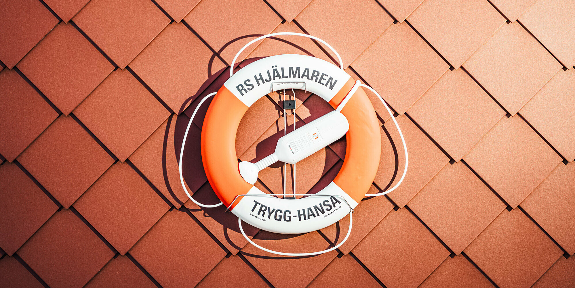 Nahaufnahme der Fassade des Seenotrettungsstationshauses am Hjälmaren See in Schweden, mit einem leuchtend orangefarbenen Rettungsring an der Wand. Der Rettungsring trägt die Aufschrift 'RS HJÄLMAREN TRYGG-HANSA', umrahmt von den charakteristischen PREFA Wandrauten 29×29 in P.10 Oxydrot, deren gleichmäßige Struktur Stärke und Zuverlässigkeit vermitteln. Der Rettungsring, ein Symbol der Sicherheit auf dem Wasser, kontrastiert eindrucksvoll mit der texturierten, roten Aluminiumoberfläche, die die Widerstandsfähigkeit des Gebäudes gegen die harten schwedischen Wetterbedingungen unterstreicht.